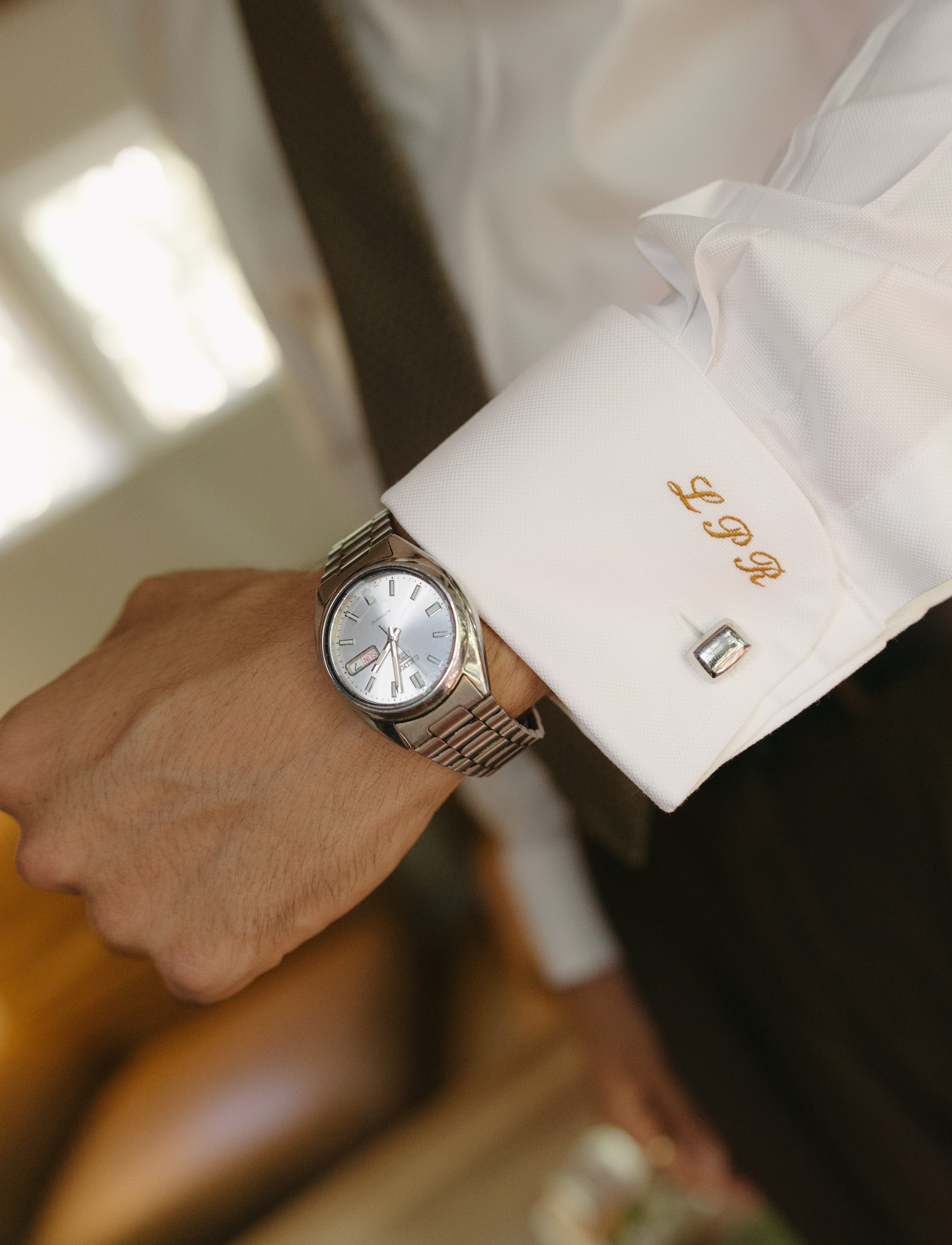 Close-up of a person's wrist showing a silver watch with a white face and a date display, with the person wearing a white dress shirt with gold embroidered initials and a silver cufflink.