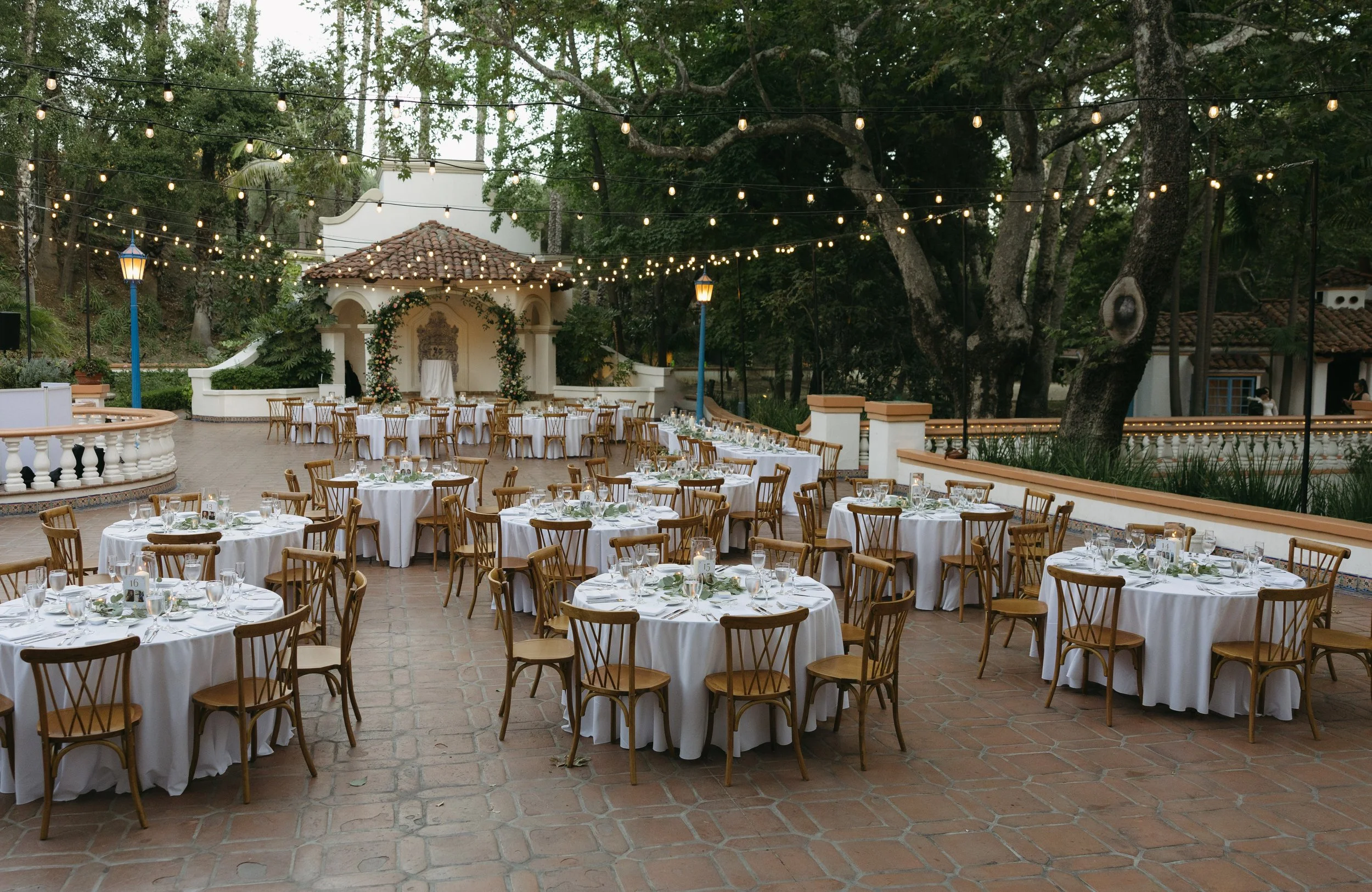 Outdoor event space with round tables covered in white tablecloths, set with glassware and silverware, surrounded by wooden chairs. String lights are overhead, and a small stage or focal point with floral decorations is at the back. Large trees and a historic building with white walls and terracotta roof tiles are in the background