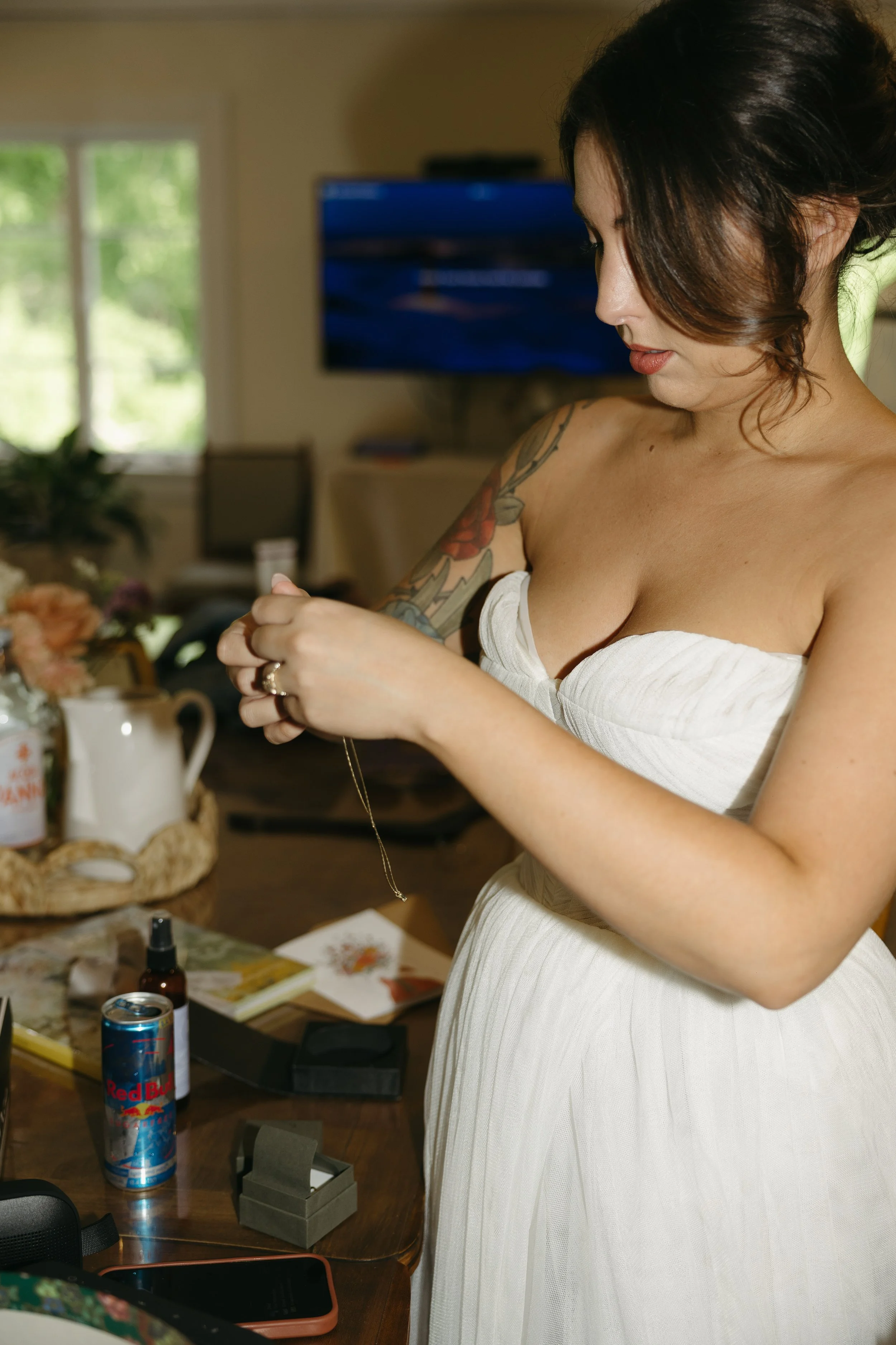 A woman in a white dress with a tattoo on her arm standing at a table, looking at a necklace she is holding.
