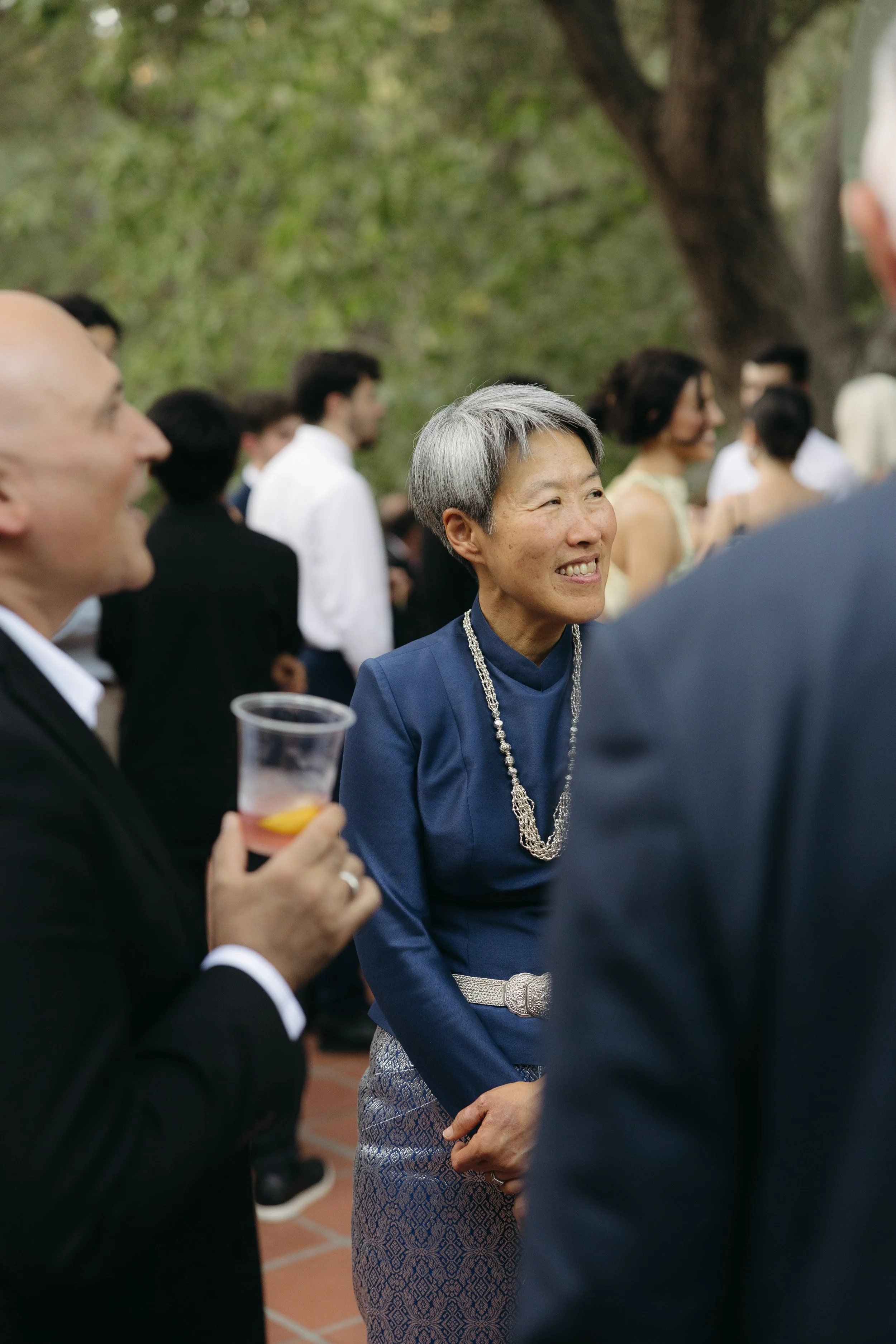 A woman with short gray hair wearing a blue top and a silver belt, smiling and engaged in conversation at an outdoor gathering, with people in formal attire mingling in the background.