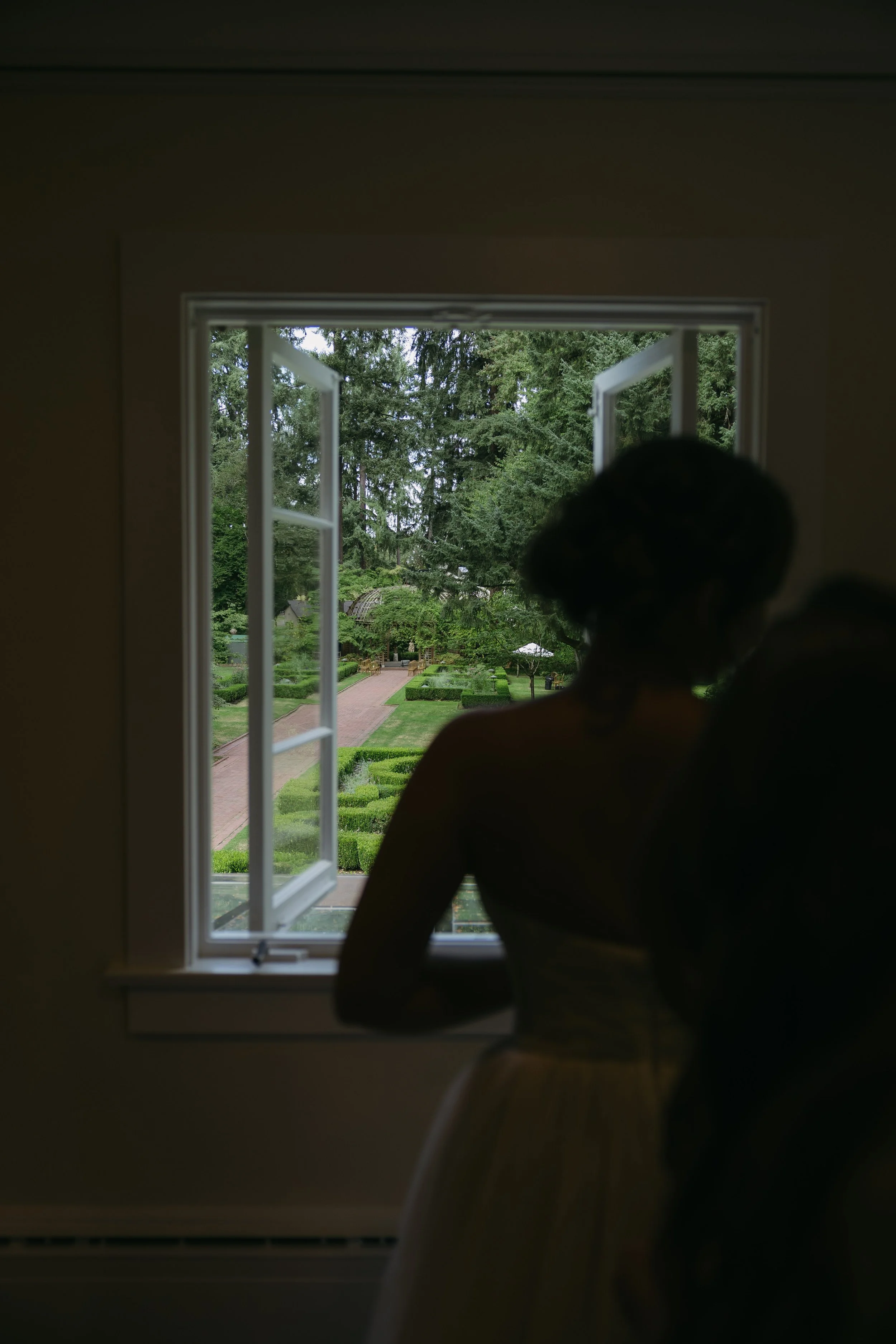 A woman in a white dress standing inside near an open window, looking outside at a lush, green garden with trees, shrubs, and a brick pathway.