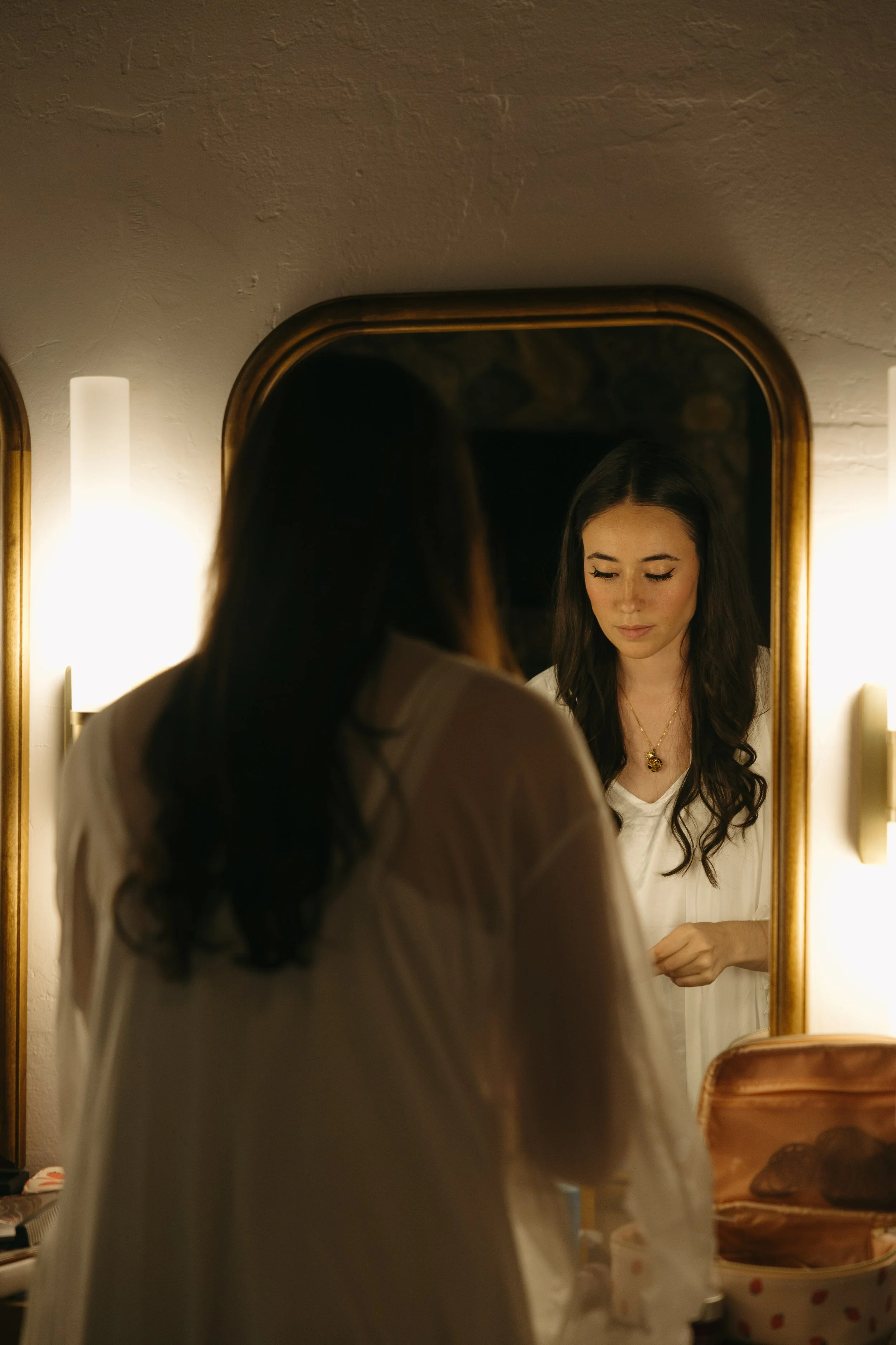 A bride with dark hair looking into a mirror, illuminated by warm lighting, wearing a white top at a Rancho Las Lomas Wedding in Los Angeles