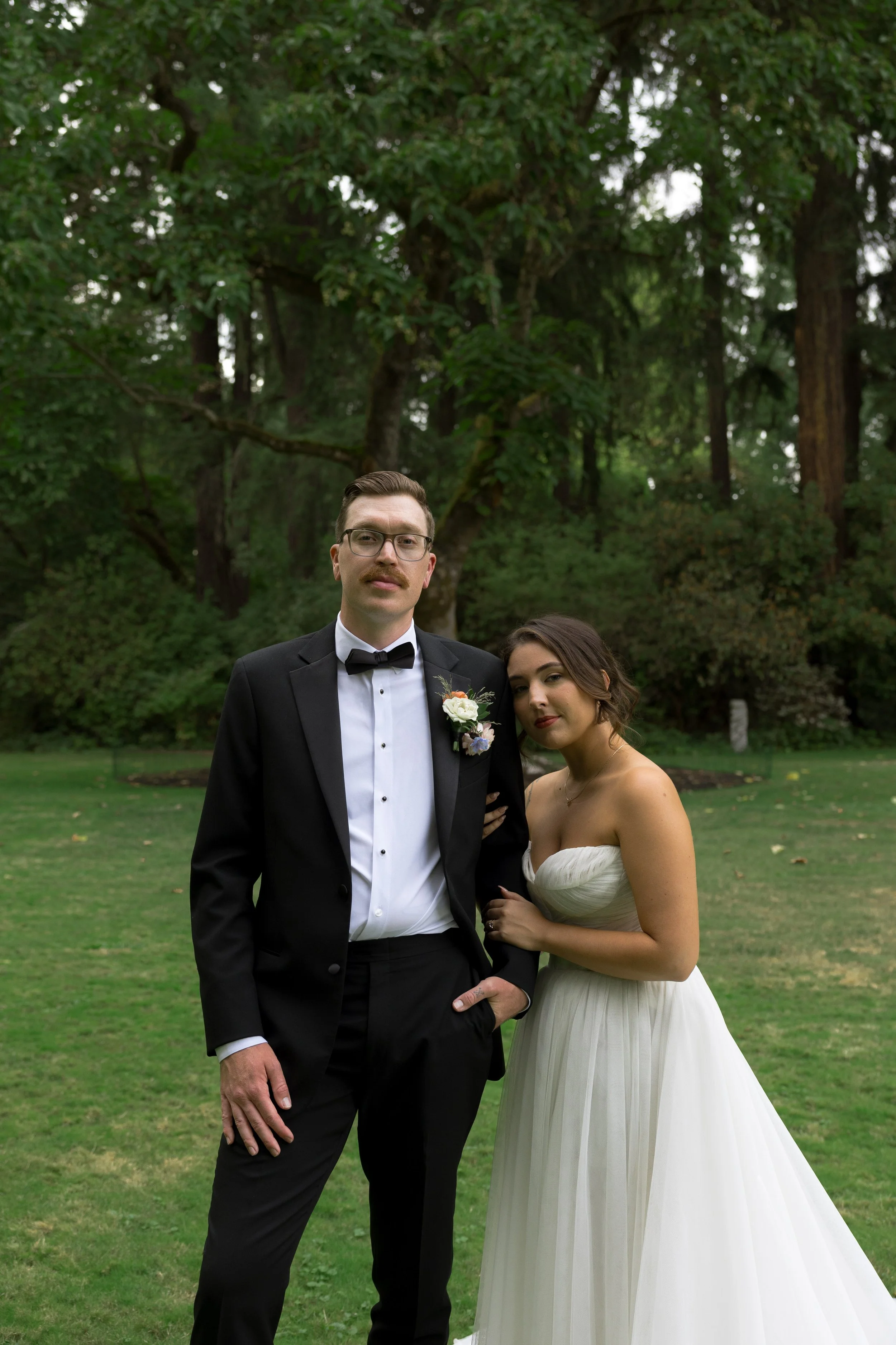 A man in a tuxedo and a woman in a white wedding dress standing outside on a grassy area with trees in the background.