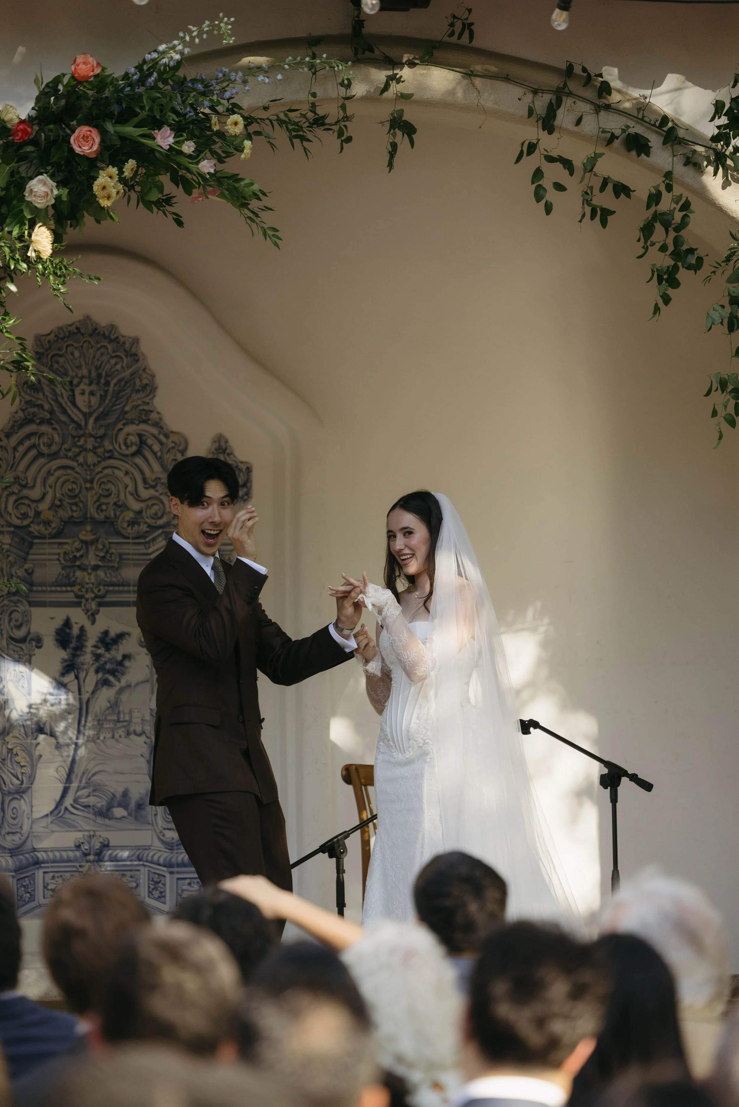 A wedding ceremony with a bride and groom smiling and holding hands on stage, audience in foreground, floral decorations overhead, and decorative wall art in the background.