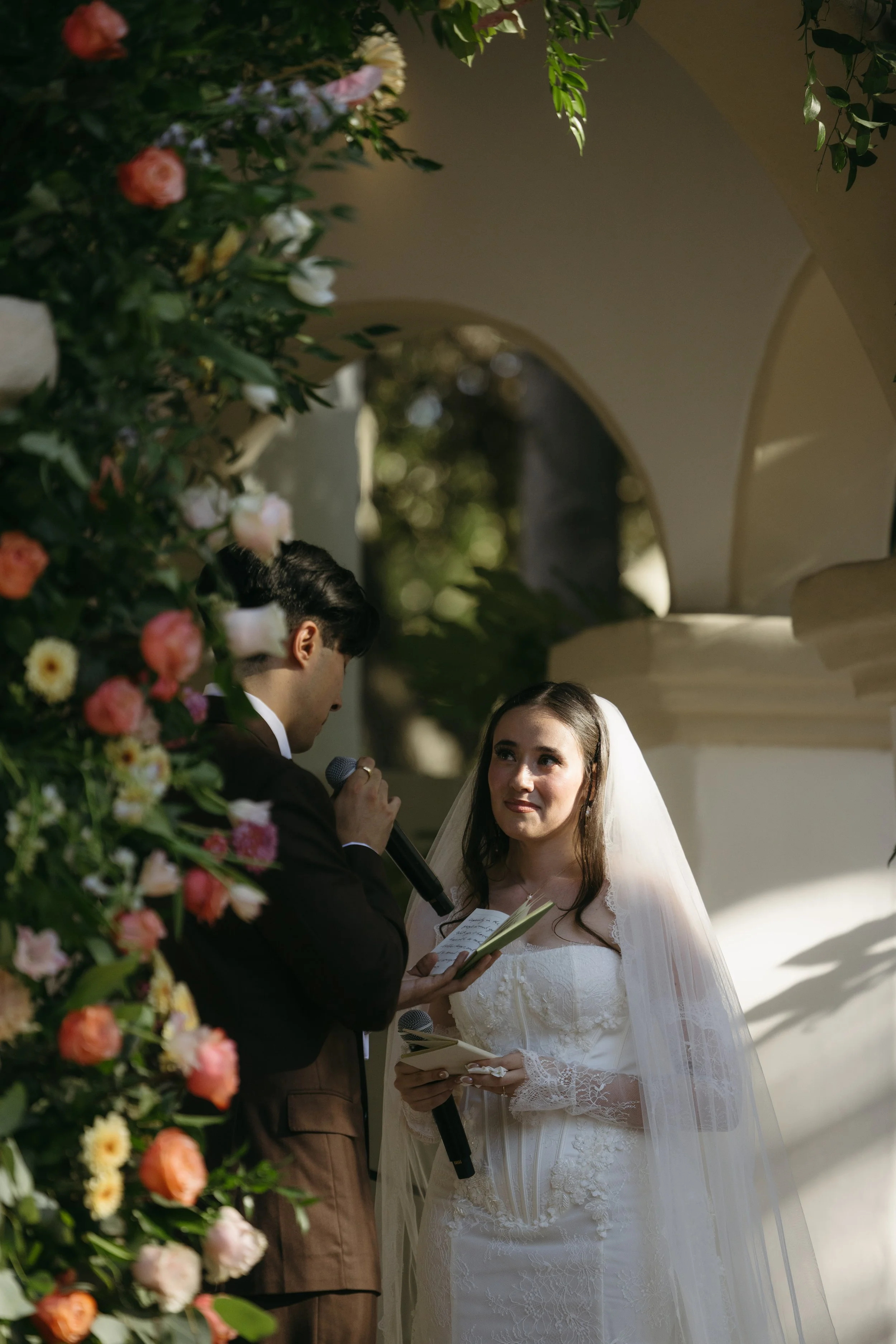A bride and groom exchange vows during their wedding ceremony outdoors, surrounded by floral decorations.