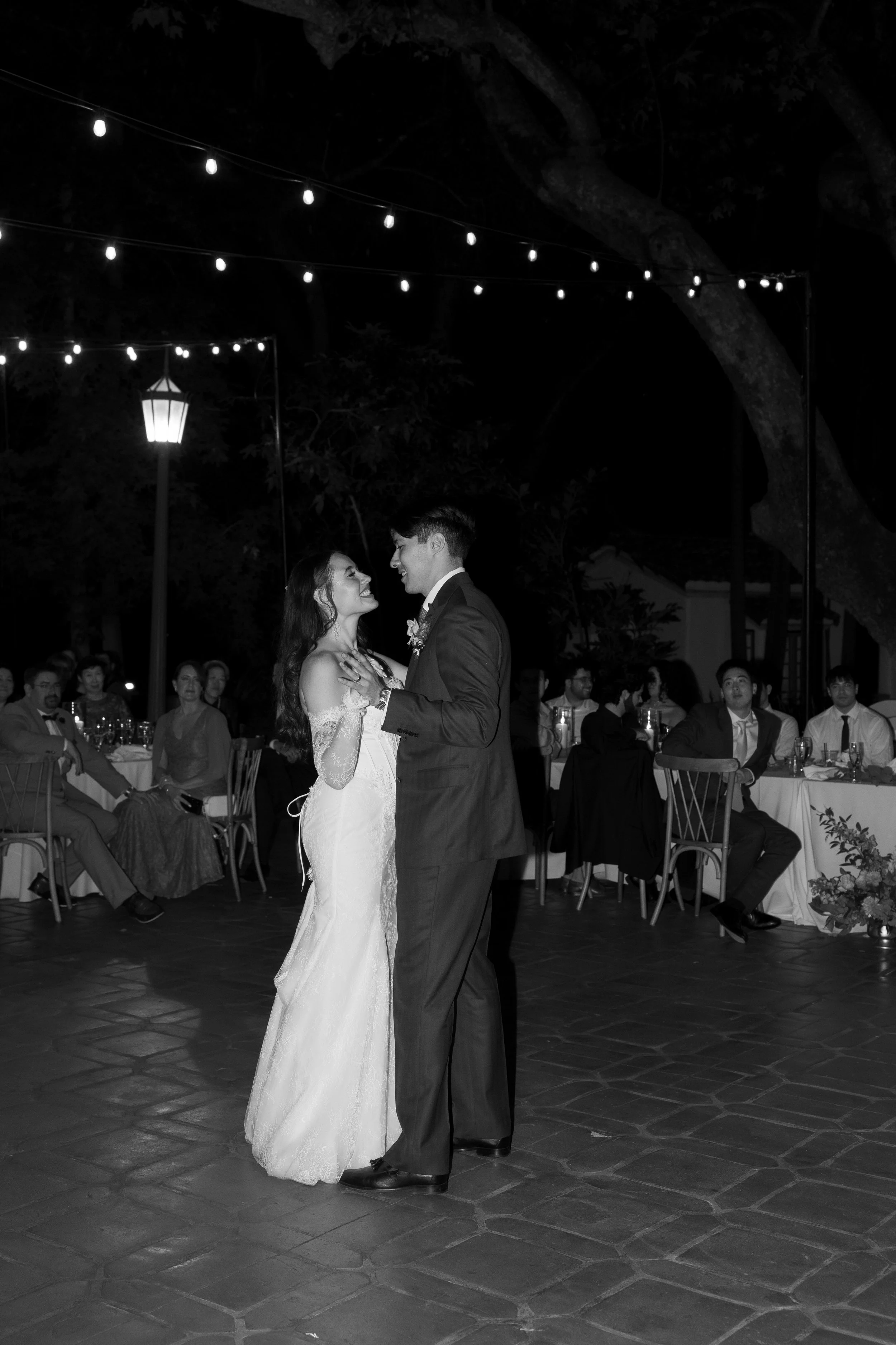 A bride and groom share a dance at their wedding reception outdoors at night, under string lights, with guests seated at tables watching.