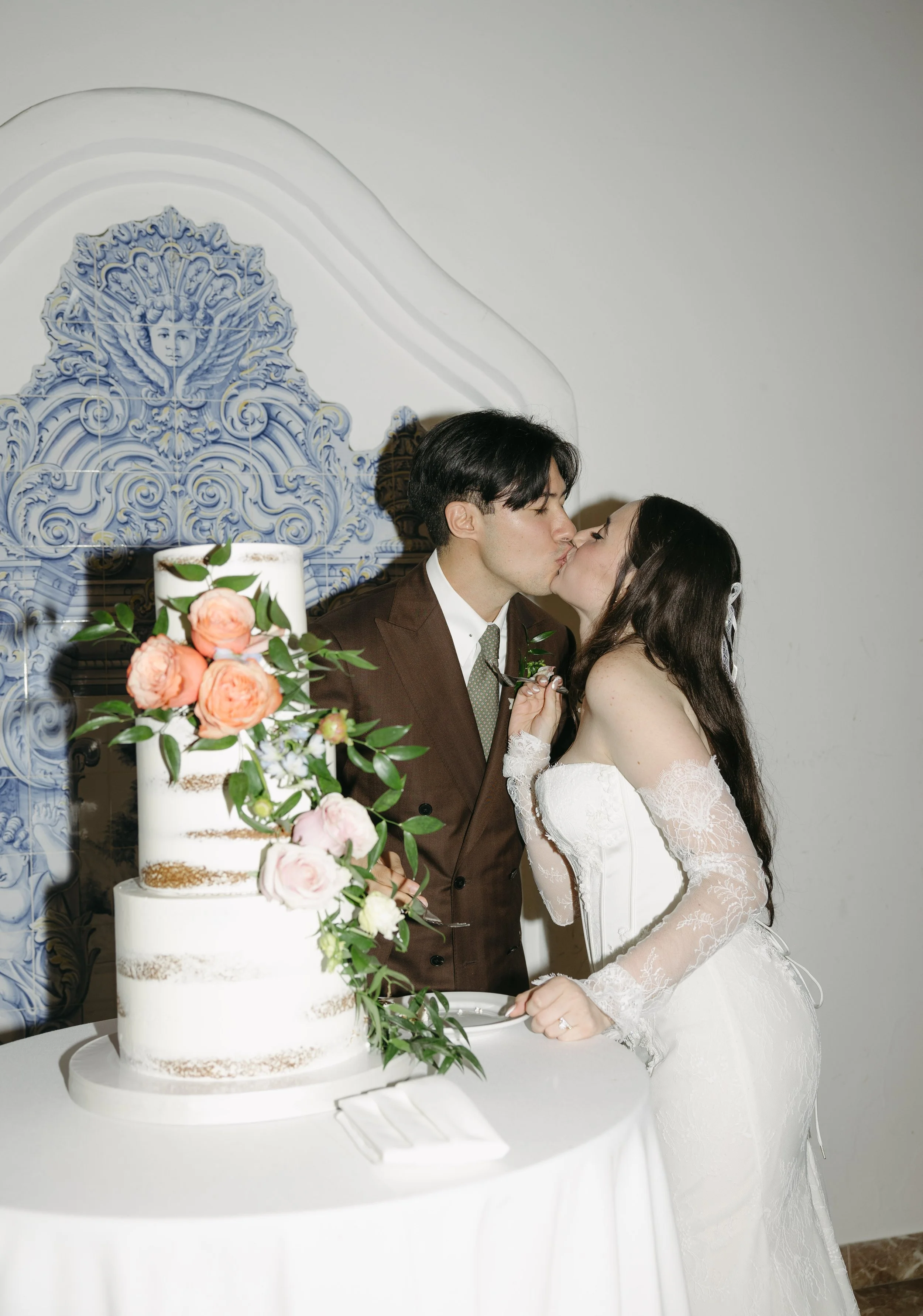 A bride and groom kiss in front of a wedding cake with pink and white roses during their wedding celebration.