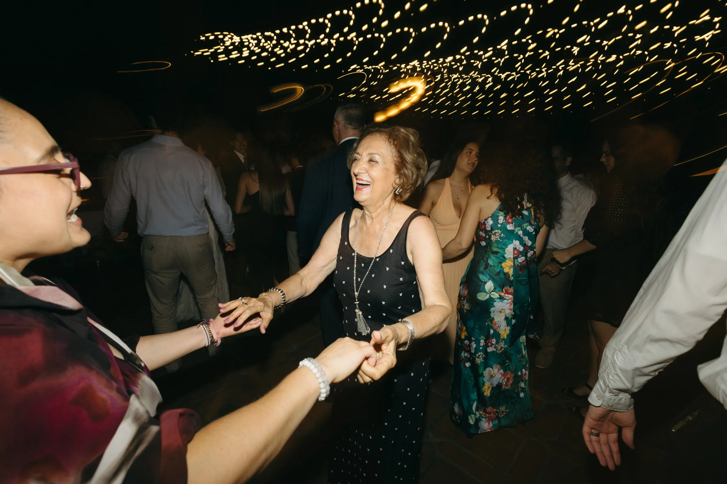 People dancing and celebrating at a party or wedding reception with string lights overhead.