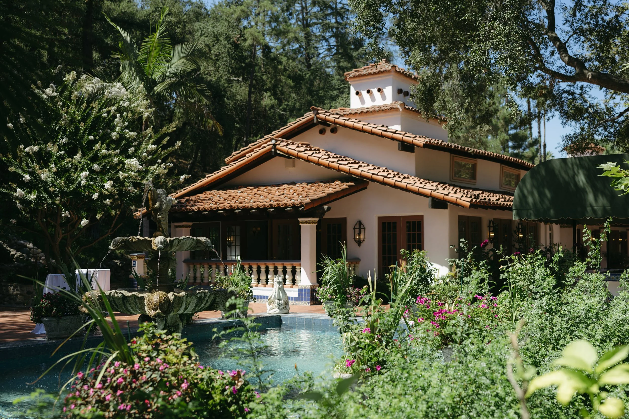 A house with Spanish-style architecture, featuring a red tile roof, surrounded by lush greenery, colorful flowers, and a pond with a fountain in the foreground at a Rancho Las Lomas Wedding in Los Angeles