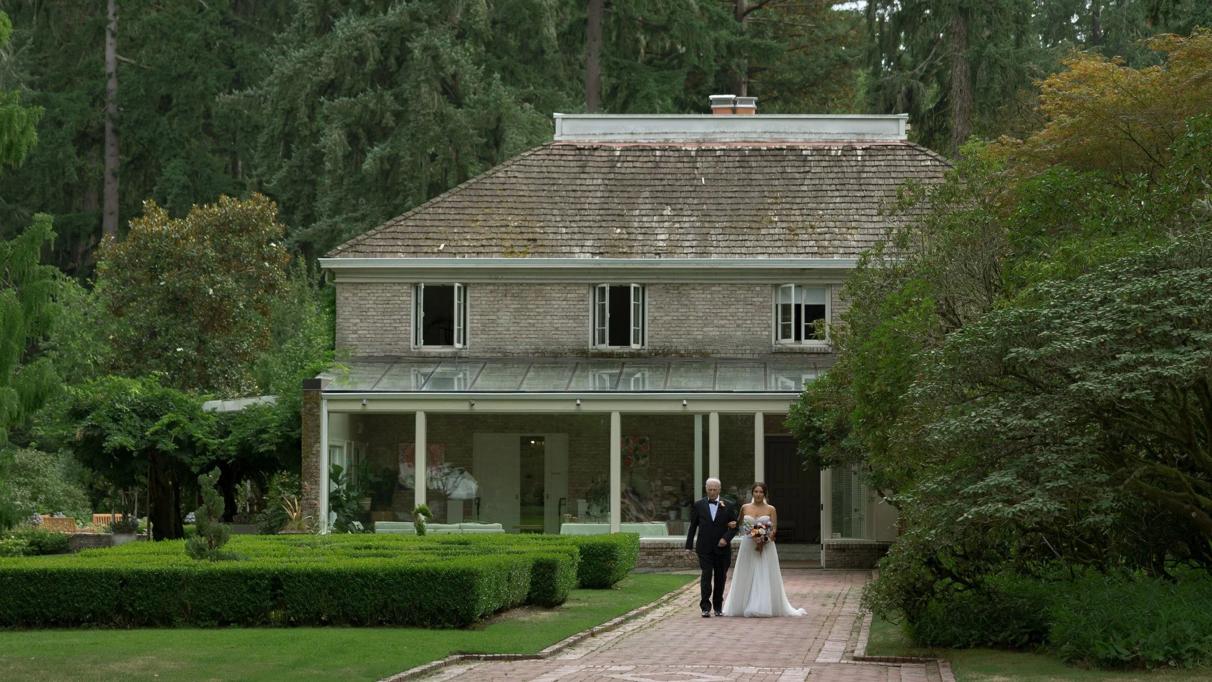 Outdoor wedding ceremony shows a bride walking down the aisle with her father, with a large Victorian house in the background at a European inspired Lakewold Gardens wedding in Seattle