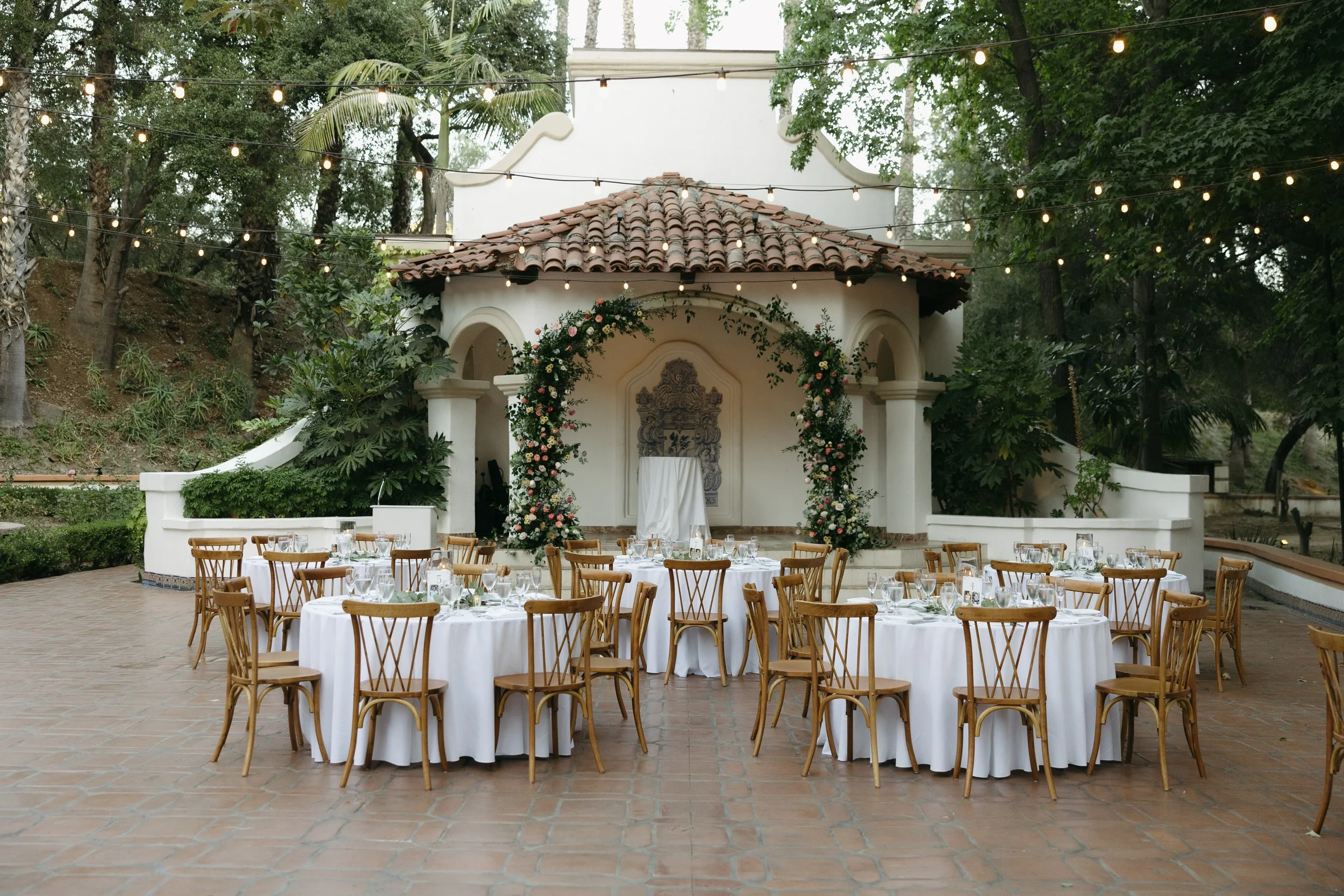 Outdoor reception decorated with glassware and centerpieces, located on a patio with a tiled roof, decorated with an arch of flowers, with string lights overhead at a Rancho Las Lomas Wedding in Los Angeles