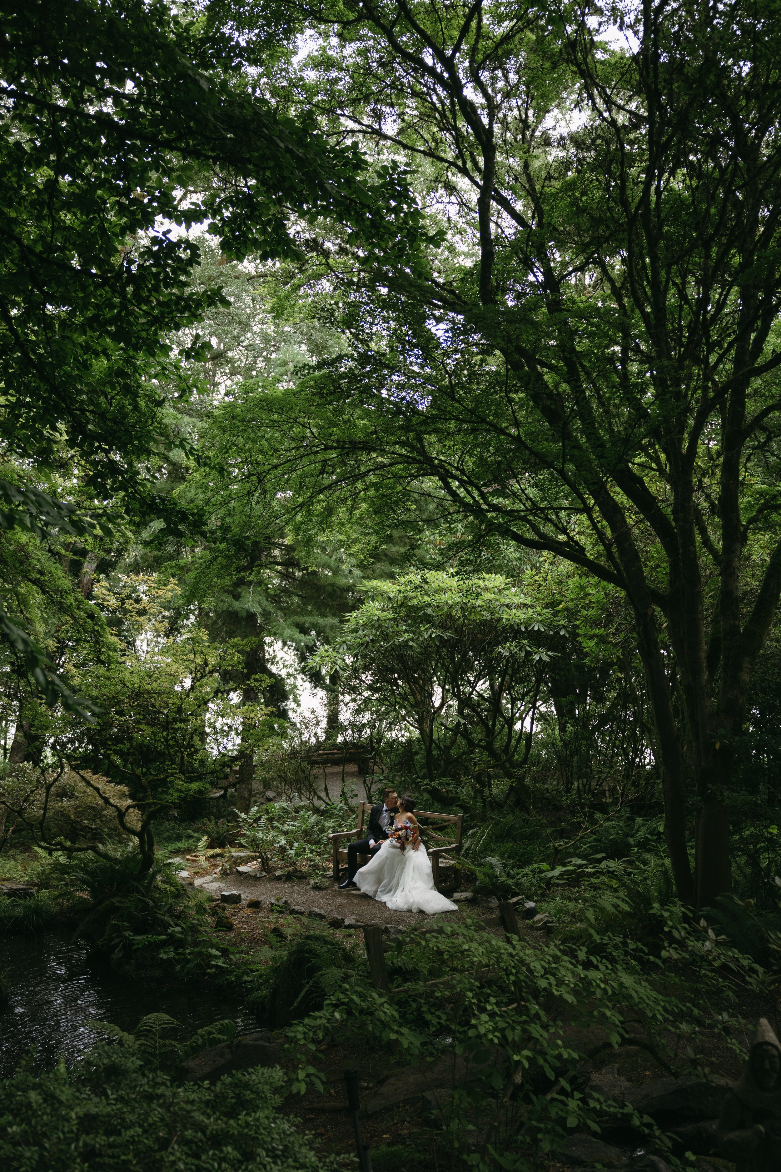 Couple in wedding attire sitting on a bench in a lush green forest, surrounded by trees and foliage at a European inspired Lakewold Gardens wedding in Seattle