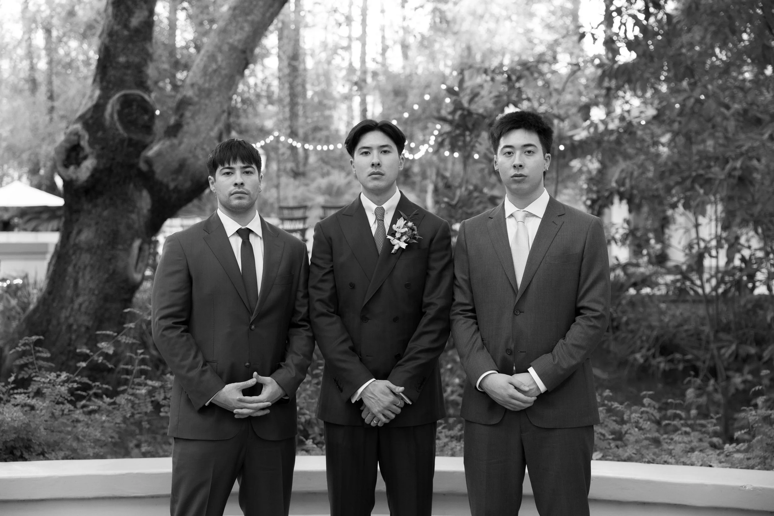 Three men dressed in suits standing outdoors in front of a large tree and string lights, with a garden in the background.