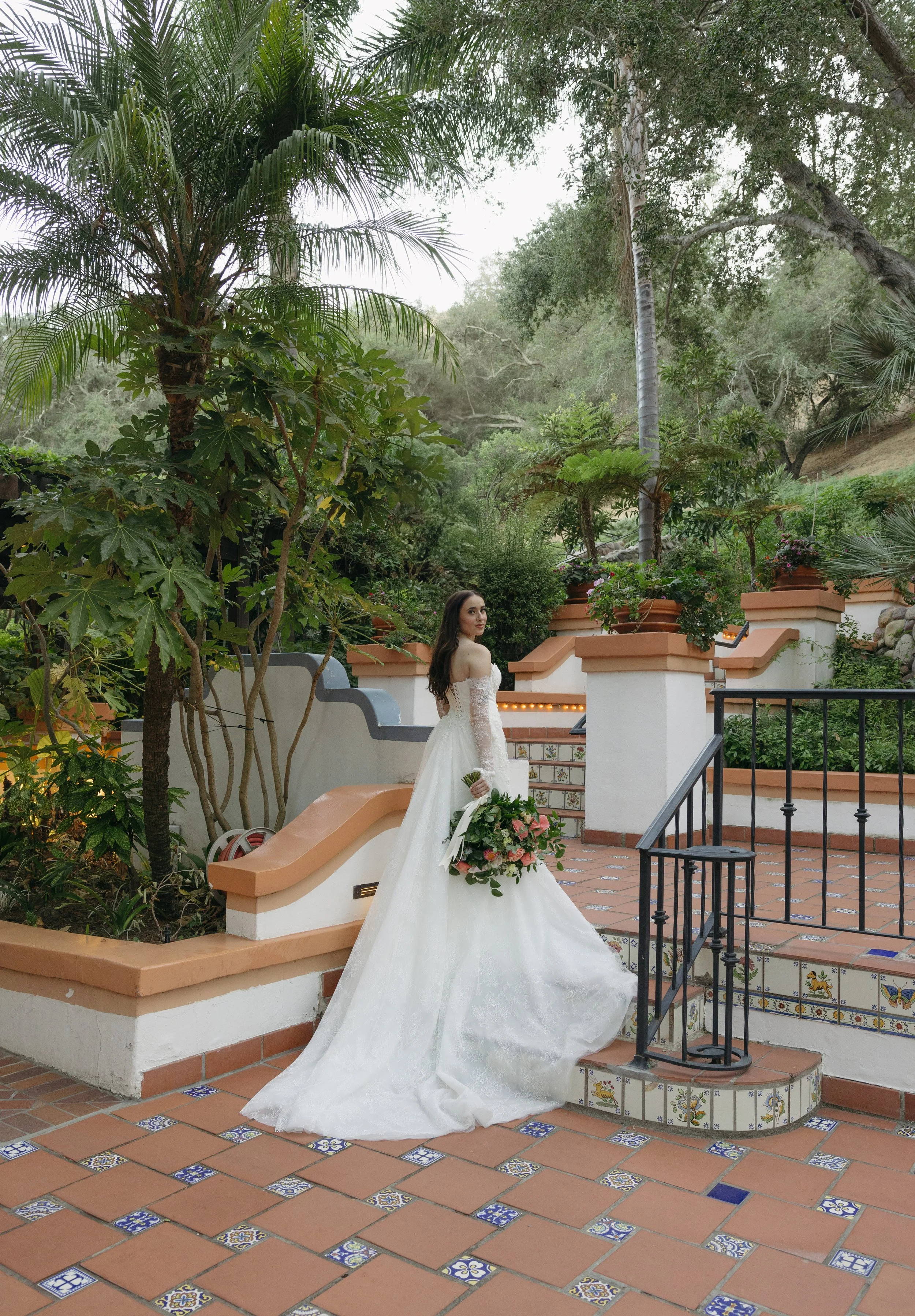 A woman in a white wedding dress holding a bouquet stands on steps in a lush garden with tall trees and potted plants at a Rancho Las Lomas Wedding in Los Angeles
