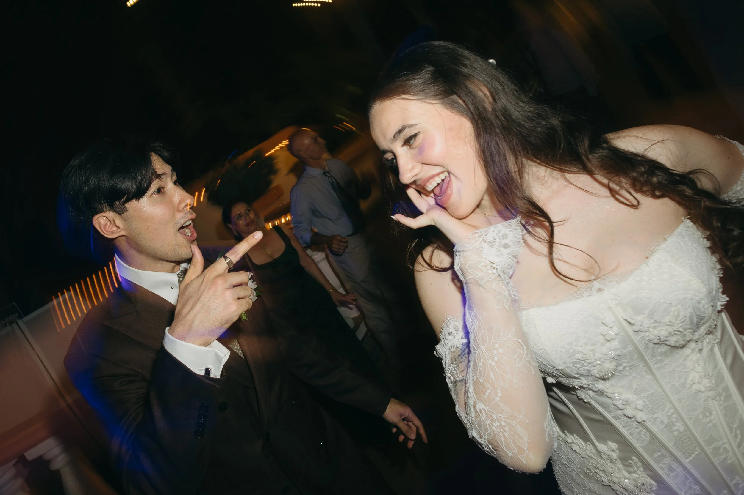 A man in a suit and a woman in a wedding dress are dancing and laughing at a wedding reception. The man appears to be pointing at the woman, and she is smiling with her eyes closed. In the background, other guests are visible, enjoying the celebratio