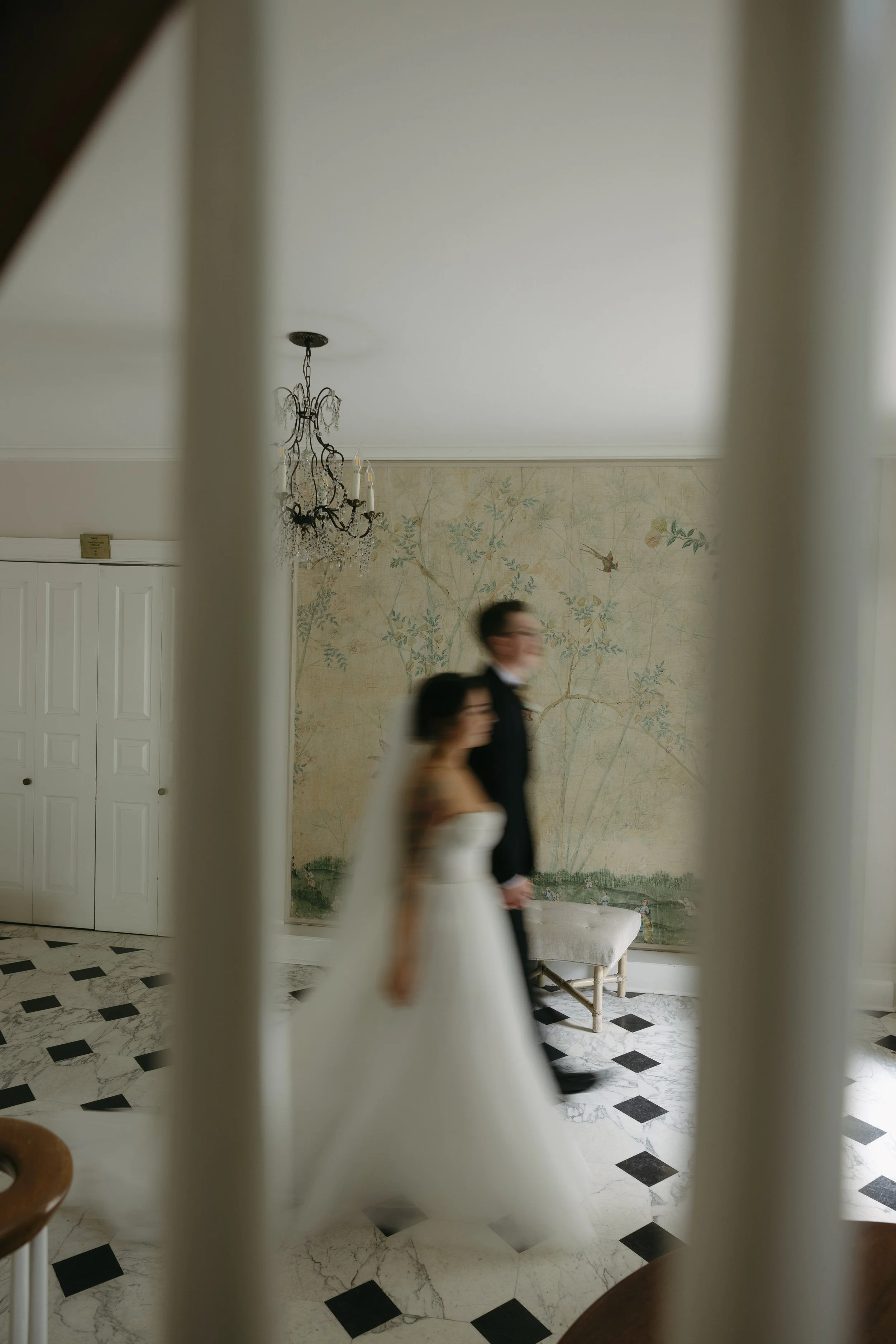 A blurry photo of a bride and groom walking inside a decorated room, seen through white vertical bars or railing. The bride wears a white wedding gown, and the groom wears a black suit. The background features a chandelier, a wall with a nature-theme