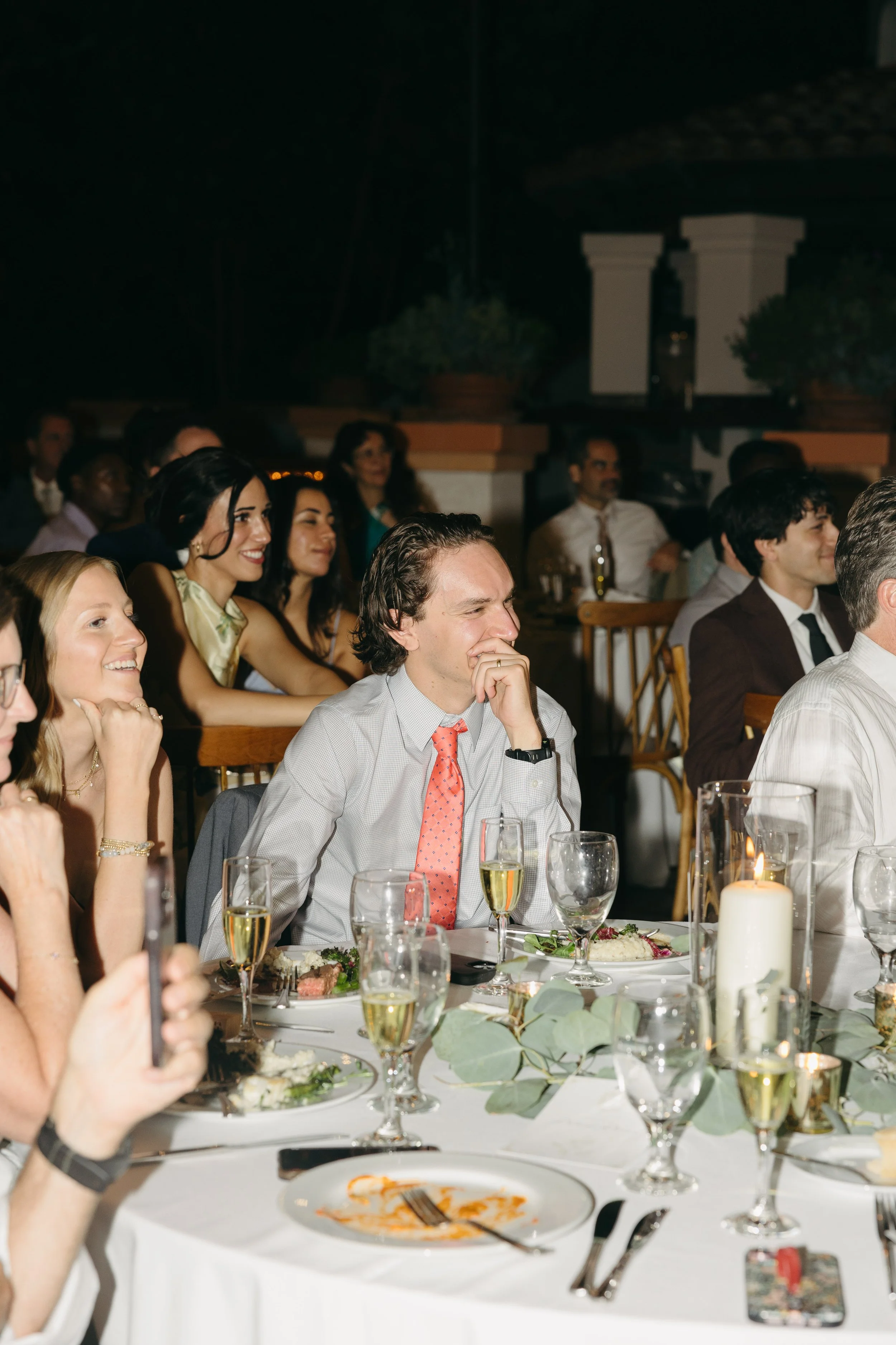 A group of people sitting at a banquet table, dressed formally, attending a celebration or wedding reception, with some people laughing and smiling.