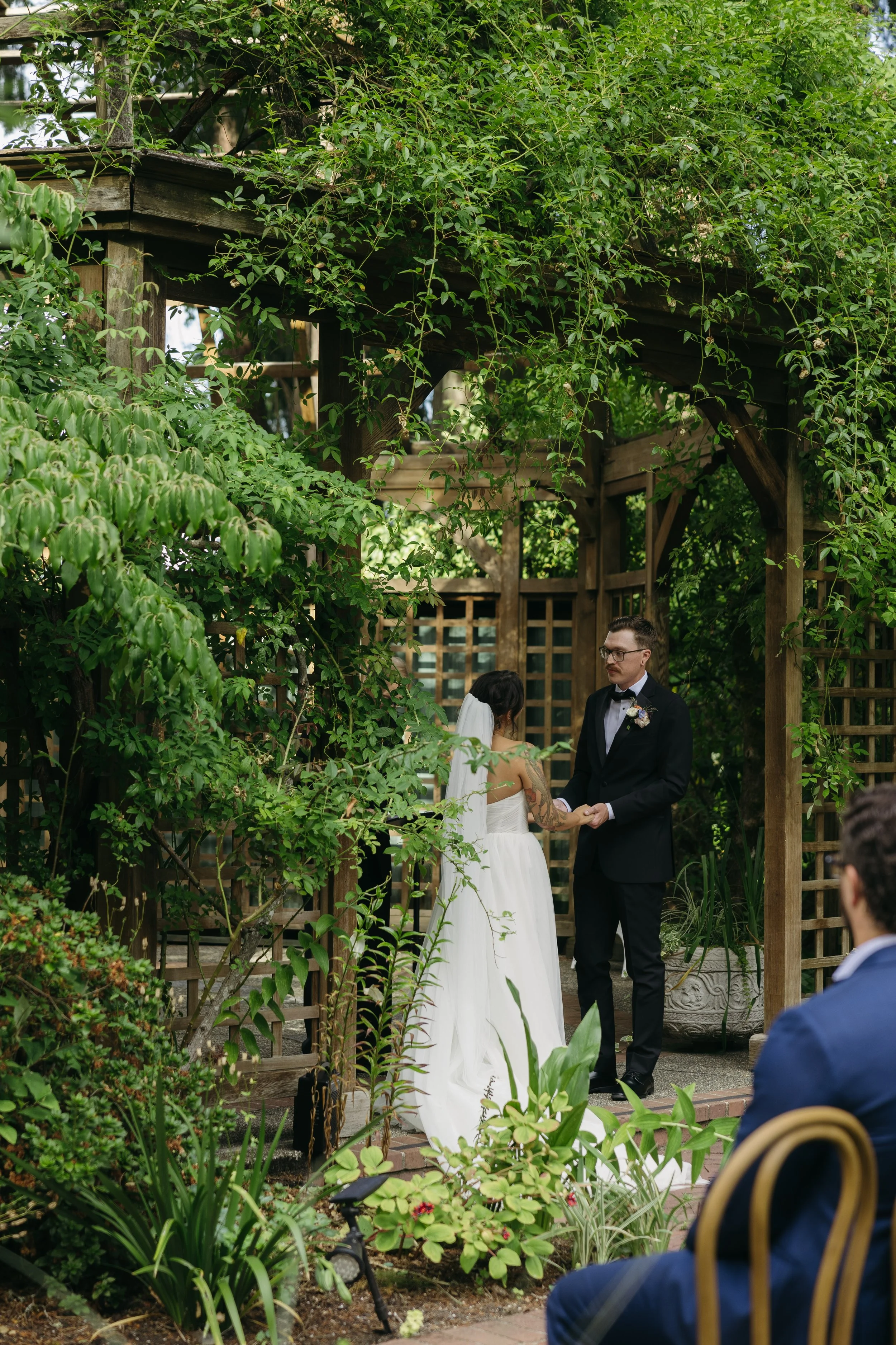 A wedding ceremony taking place outdoors under a leafy green arbor, with a bride and groom holding hands, surrounded by greenery and garden decor.