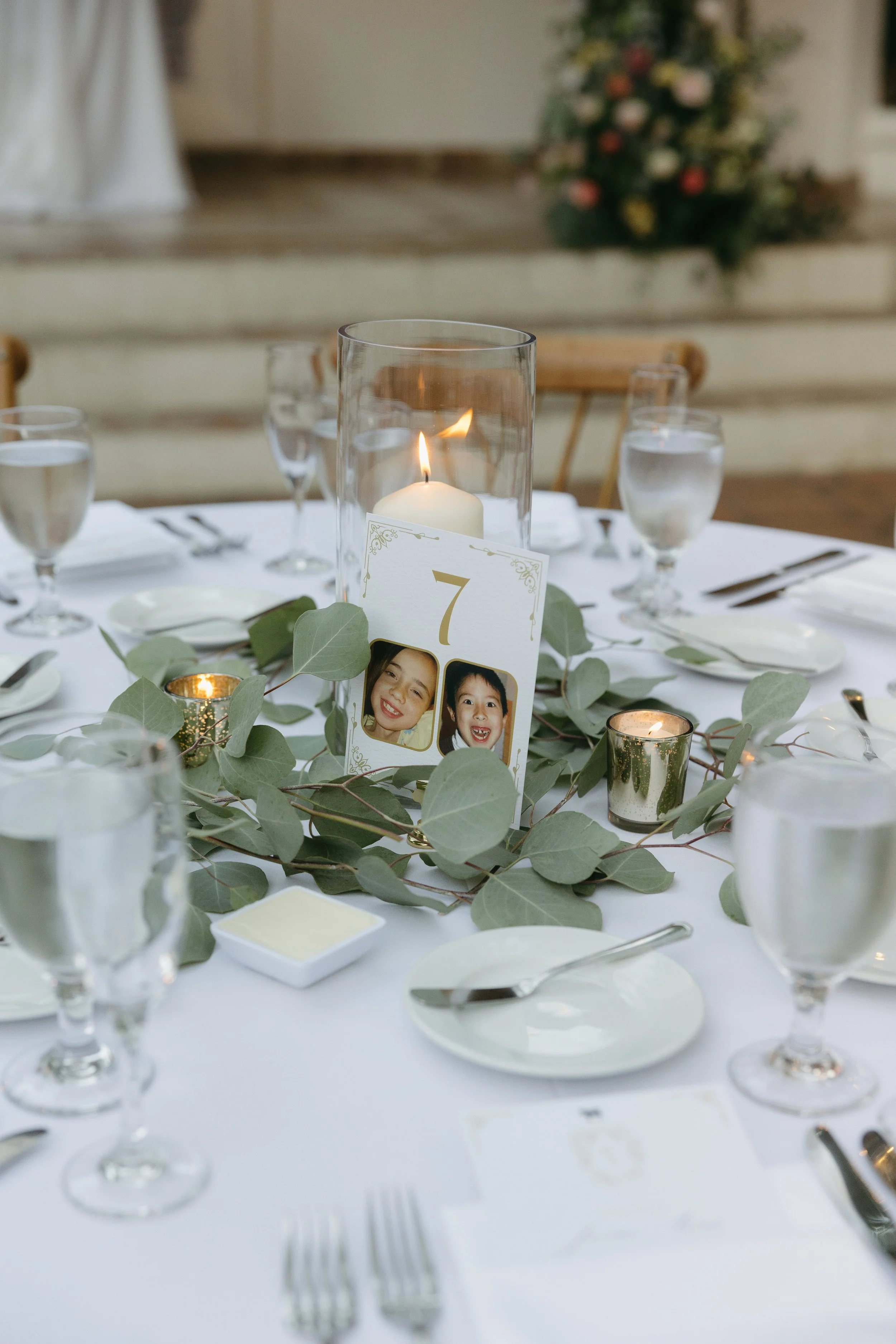 Table centerpiece with a candle in a glass holder, surrounded by greenery and small gold candles, with a place card displaying the number 7 and photos of two children, set for a formal event.