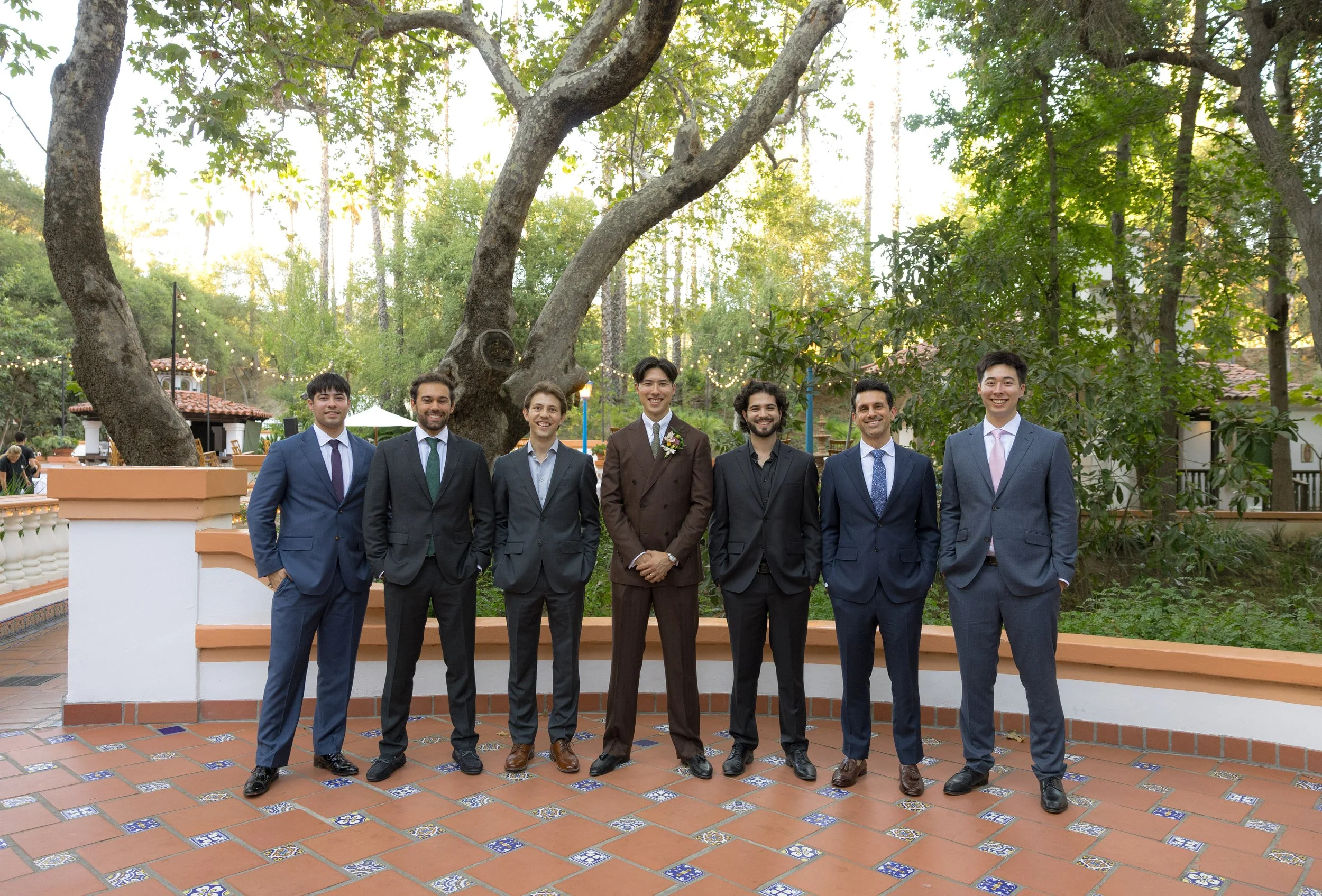 Seven men in suits standing outdoors in front of large trees and a decorative wall, smiling for the camera.