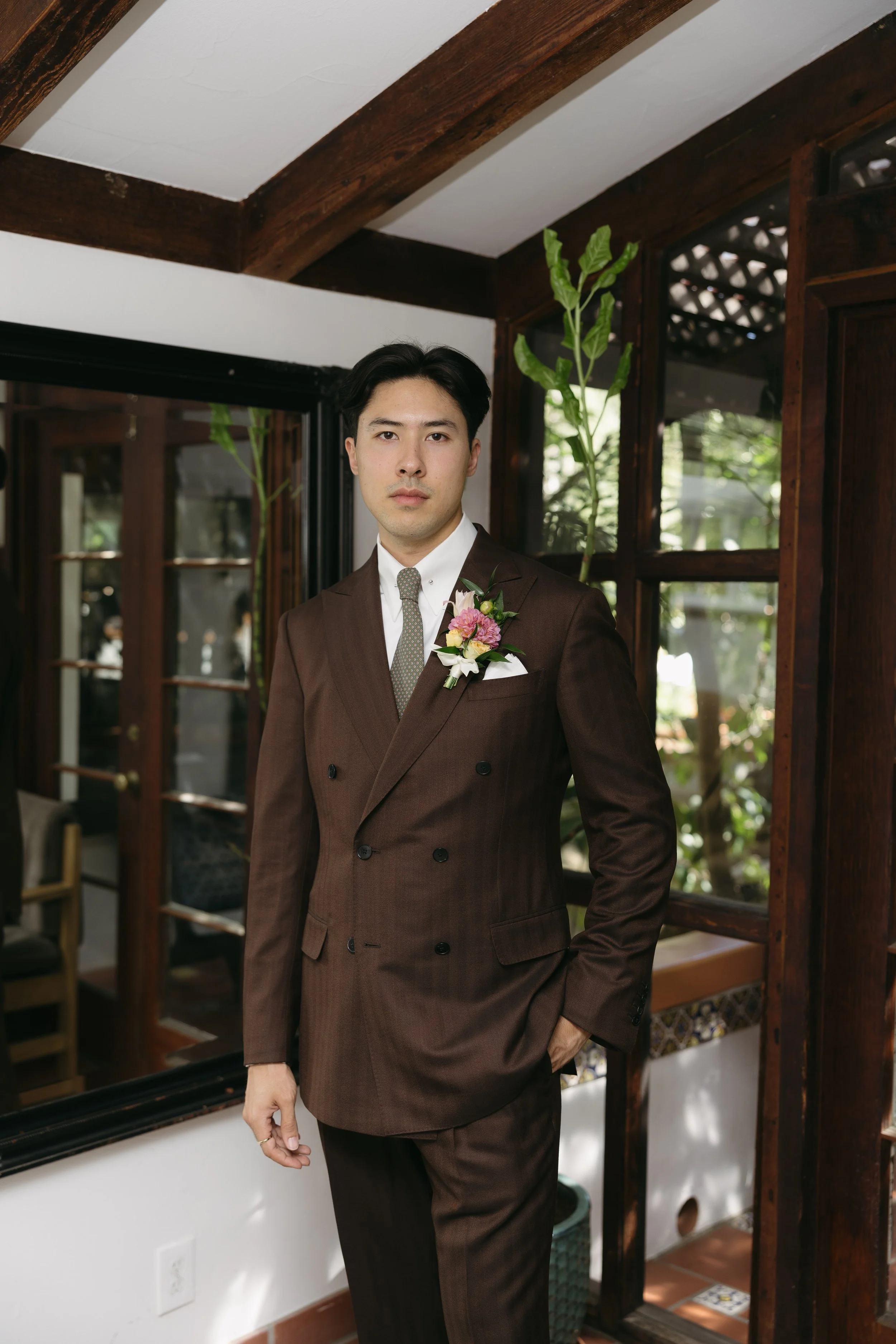 Man in a brown suit with a boutonnière, standing indoors near a window with wooden framing and plants in the background.