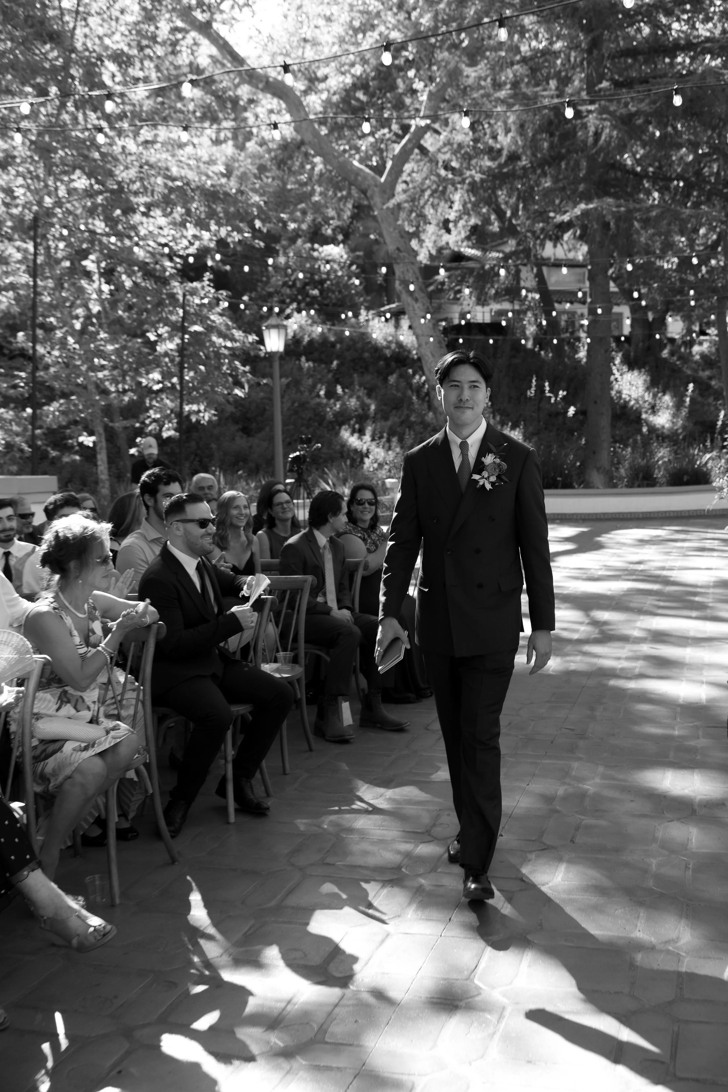 A man in a black suit walking towards the camera at an outdoor wedding ceremony, with guests seated on either side clapping and smiling, under string lights hanging from trees in a garden setting.