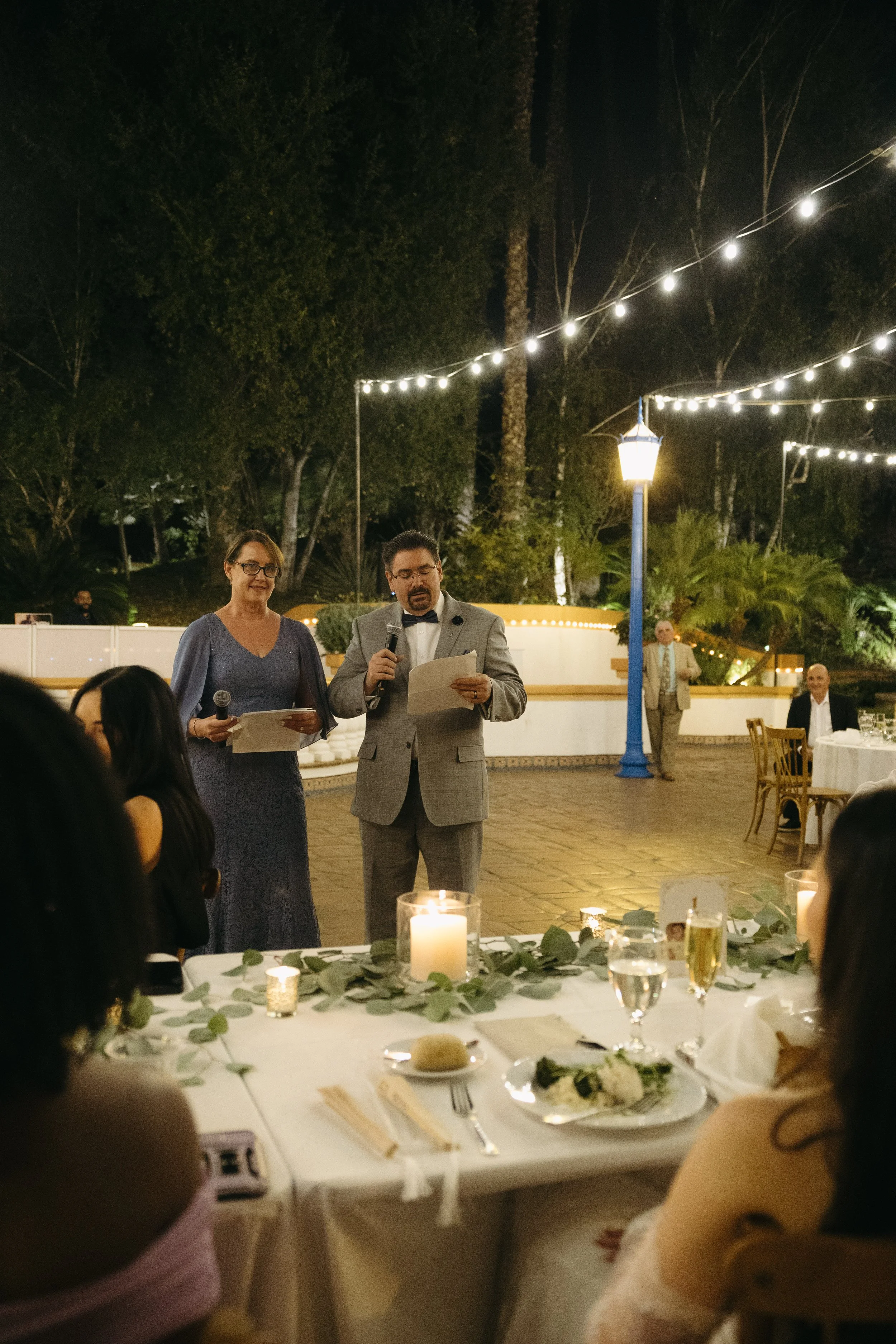 A man in a gray suit and a woman in a purple dress are giving speeches at an outdoor nighttime wedding reception, with guests seated at tables decorated with candles and greenery.