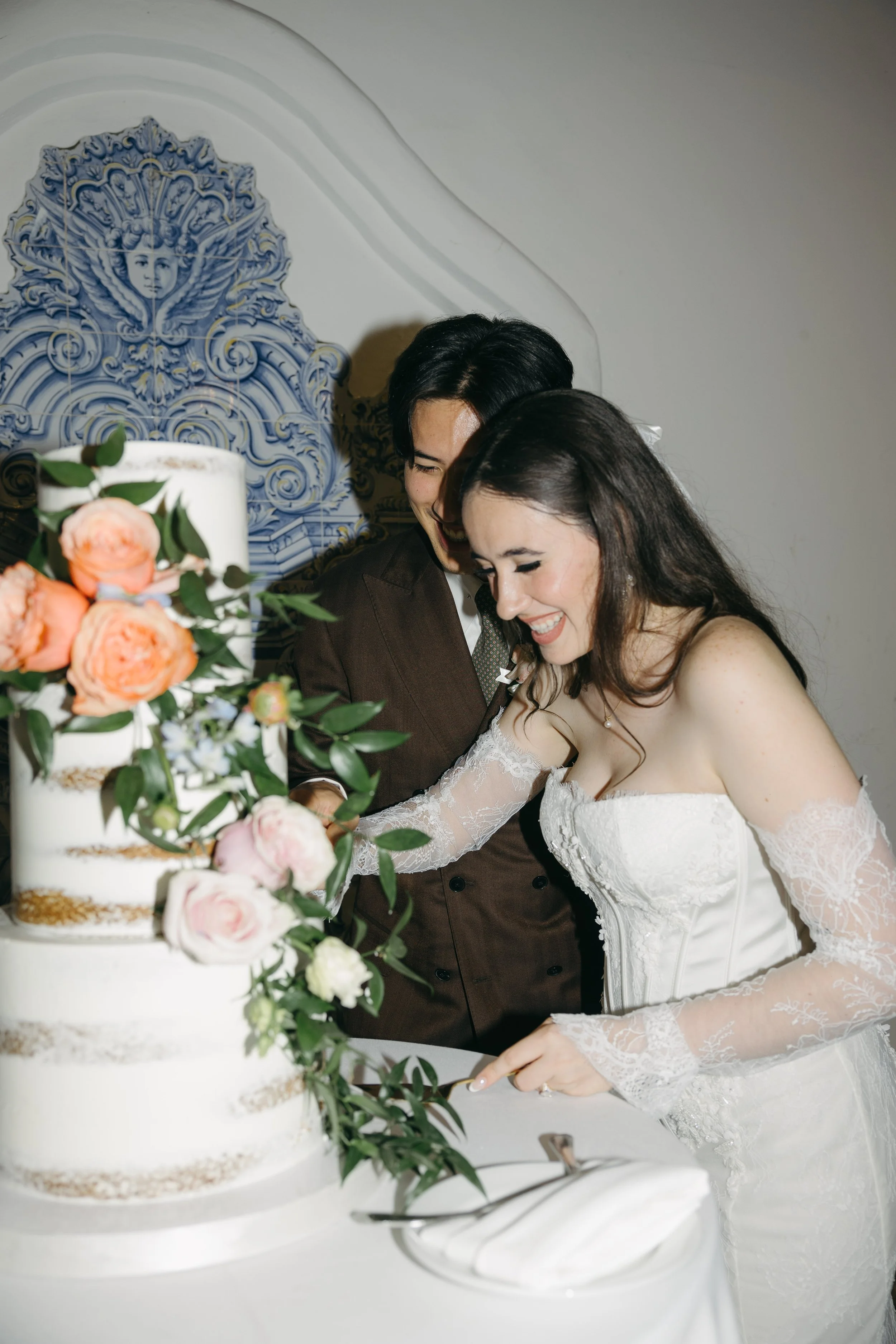 A bride and groom smiling as they cut a wedding cake decorated with peach and pink flowers and green leaves.