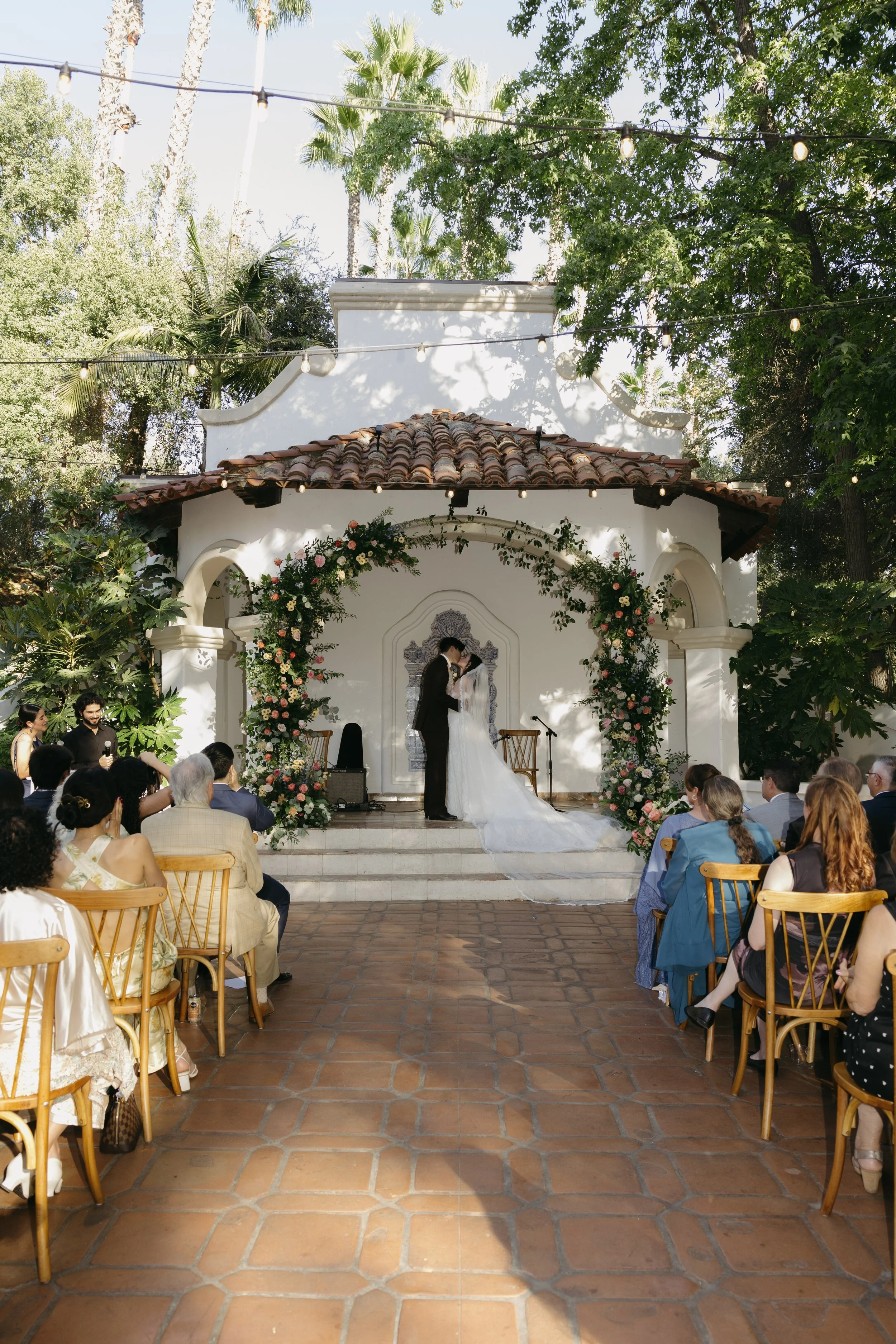 A wedding ceremony taking place outdoors under a tiled roof. The bride and groom have their first kiss, there are string lights overhead amidst trees and greenery at a Rancho Las Lomas Wedding in Los Angeles