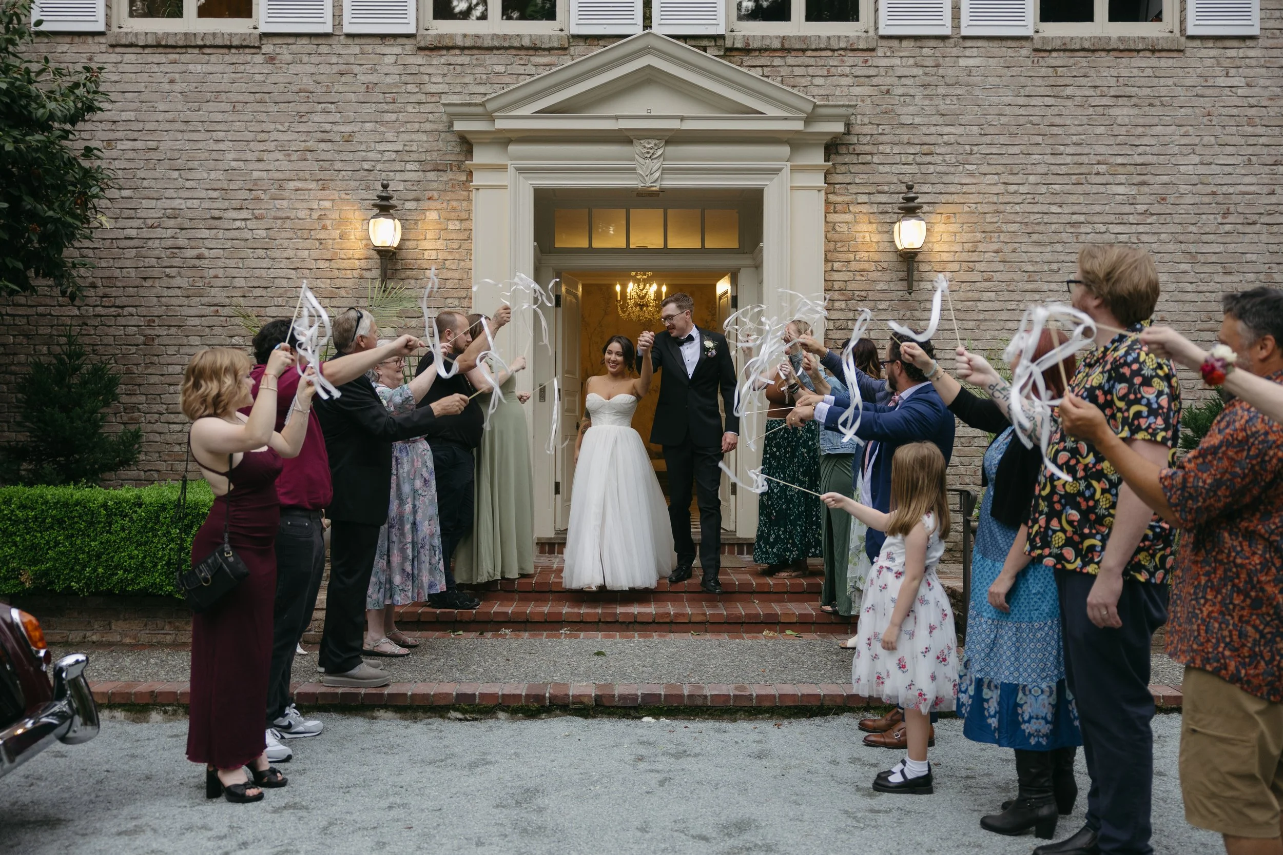 Celebrating newlyweds outside a brick building, with guests holding ribbons and cheering during their sendoff at a European inspired Lakewold Gardens wedding in Seattle