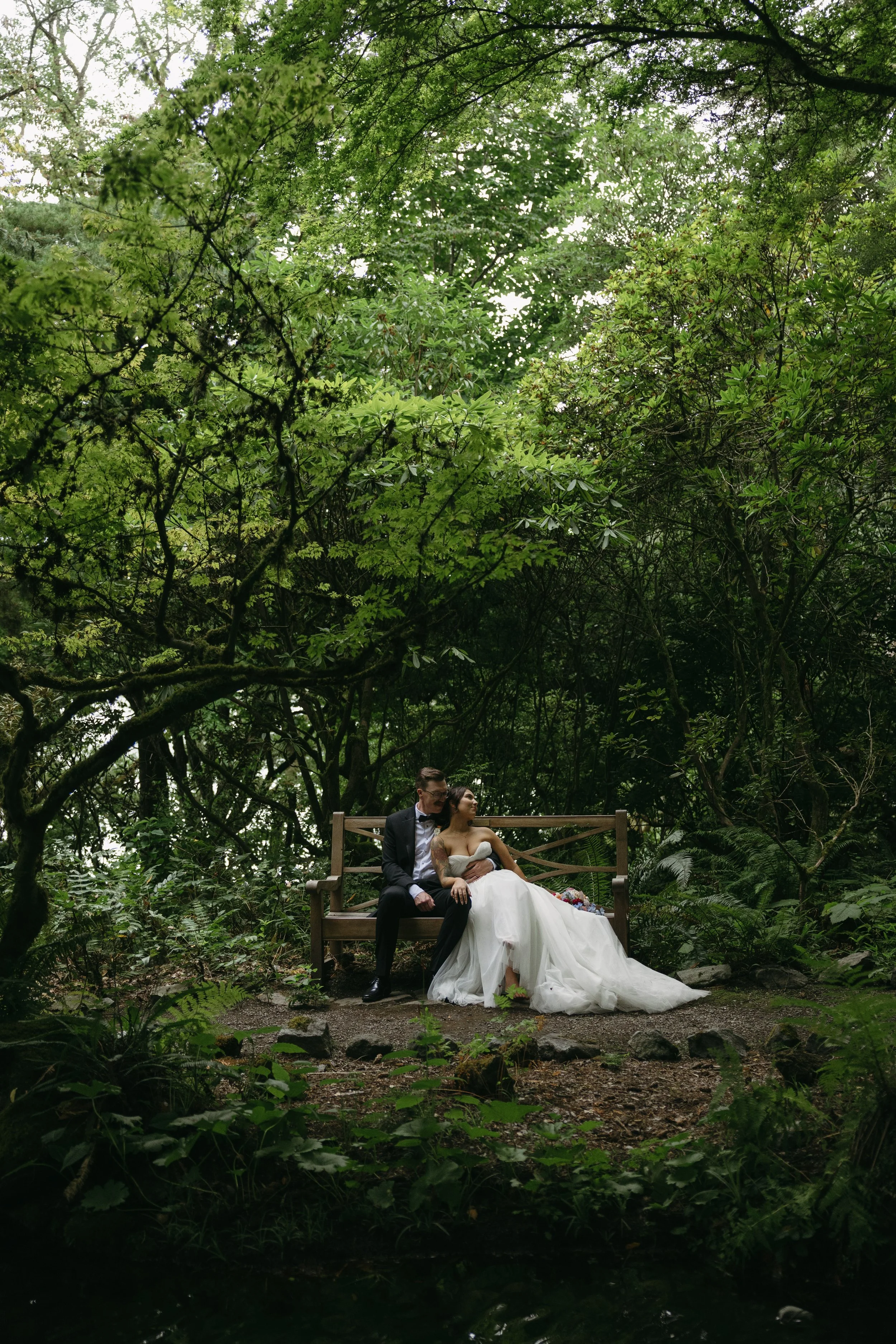 A bride and groom sitting on a wooden bench in a lush, green, forested area, sharing an intimate moment, with the bride in a white wedding gown and the groom in a tuxedo at a European inspired Lakewold Gardens wedding in Seattle