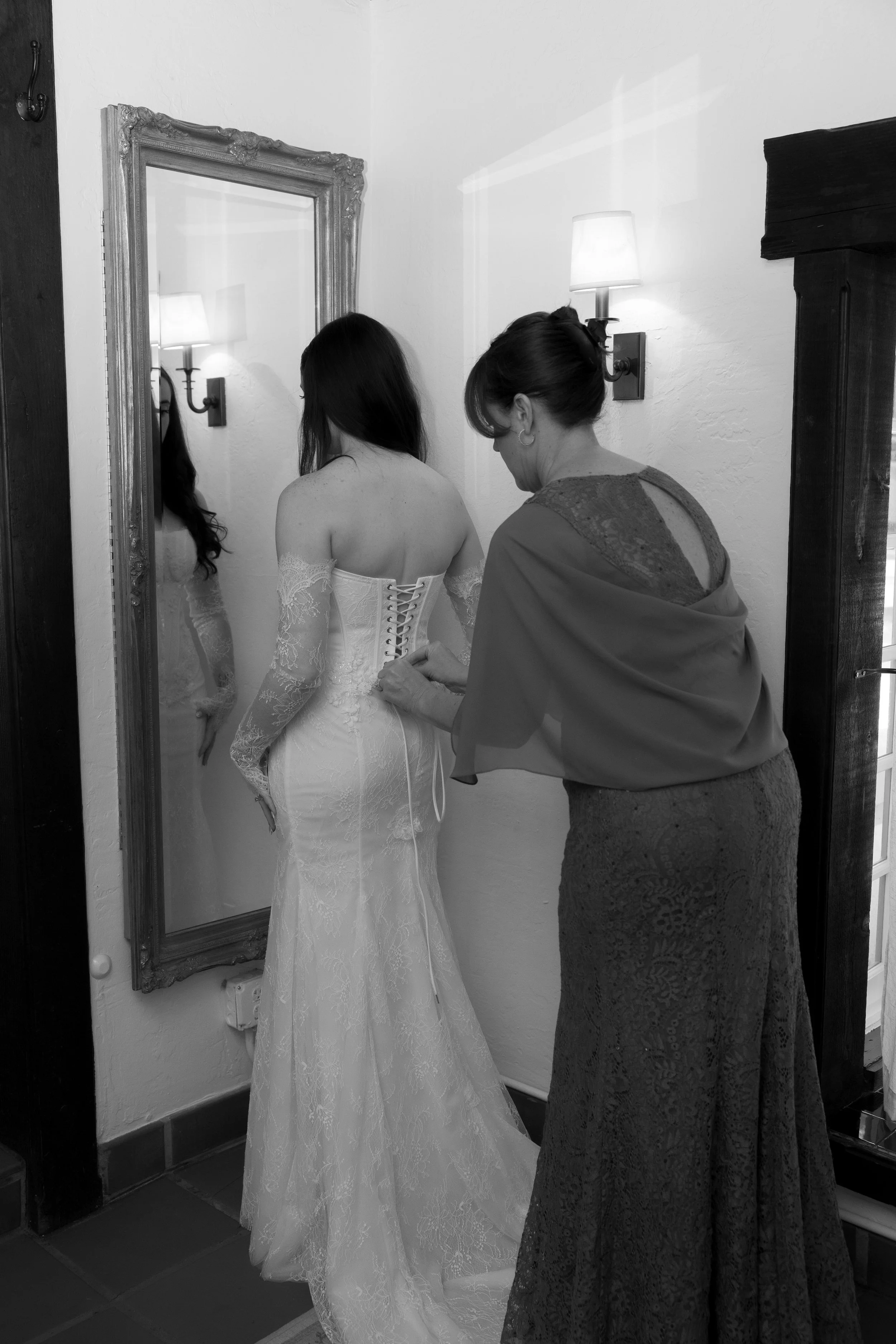 A woman in a wedding dress is being assisted with lacing it up by another woman in a long skirt and shawl, in front of a mirror in a room with wall-mounted lamps.