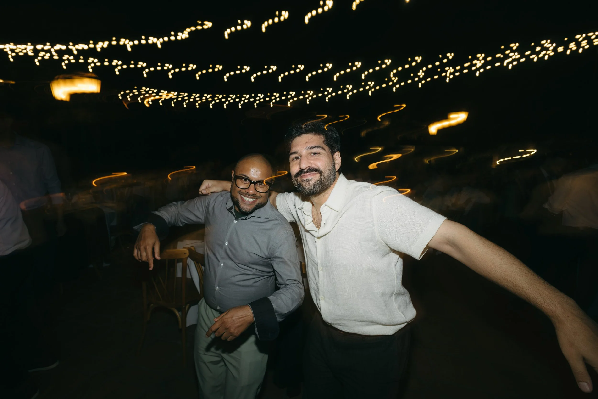 Two men are smiling and posing together at a party or celebration under string lights, with a dark background and blurred lights creating a festive atmosphere.
