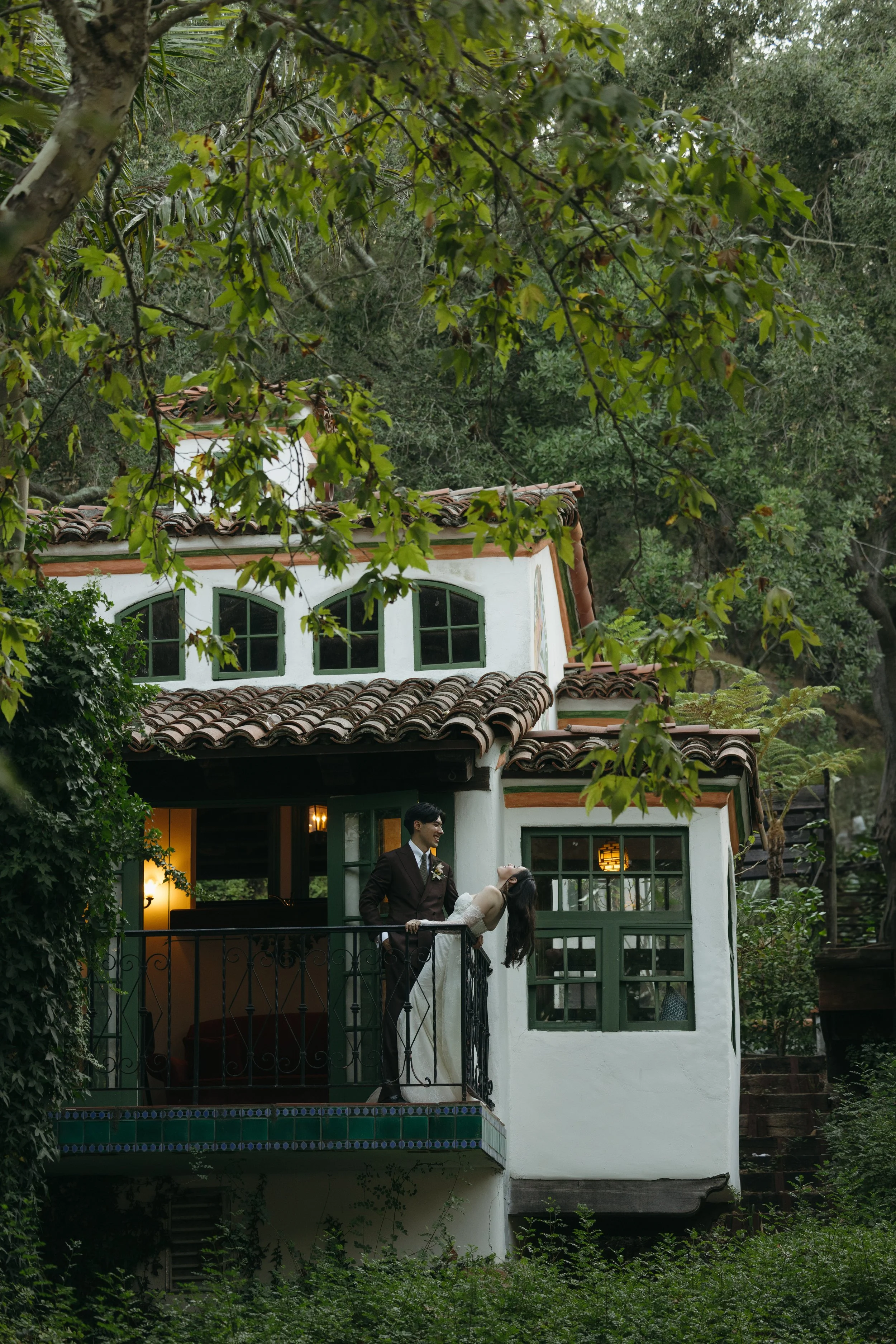 A newlywed couple on a balcony of a white house with green window frames, surrounded by green trees. The groom is dressed in a dark suit, holding the bride who is in a white wedding gown, leaning back and smiling at a Rancho Las Lomas Wedding in Los 
