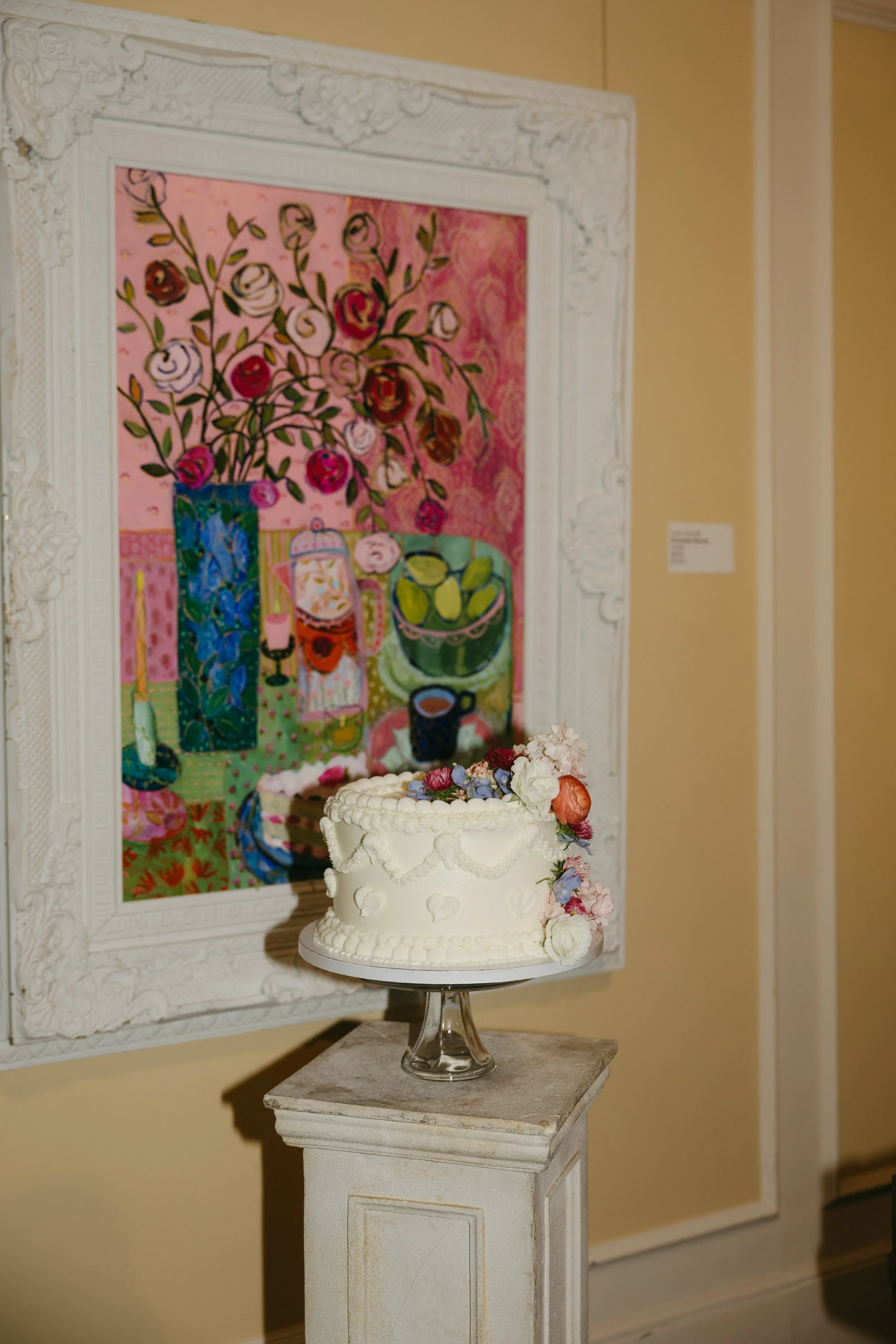 A white wedding cake decorated with flowers on a glass cake stand, placed on a white pedestal, with a colorful still life painting of a vase with flowers, fruit, and a pitcher hanging on the wall behind it.