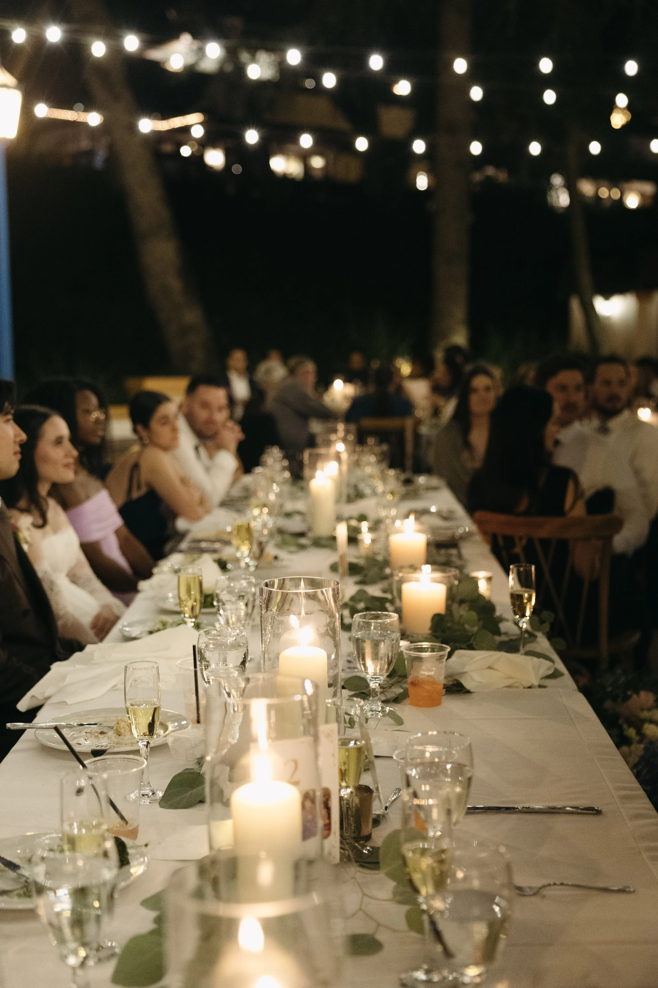 A long dinner table decorated with candles and greenery at night, surrounded by guests outdoors with string lights overhead.