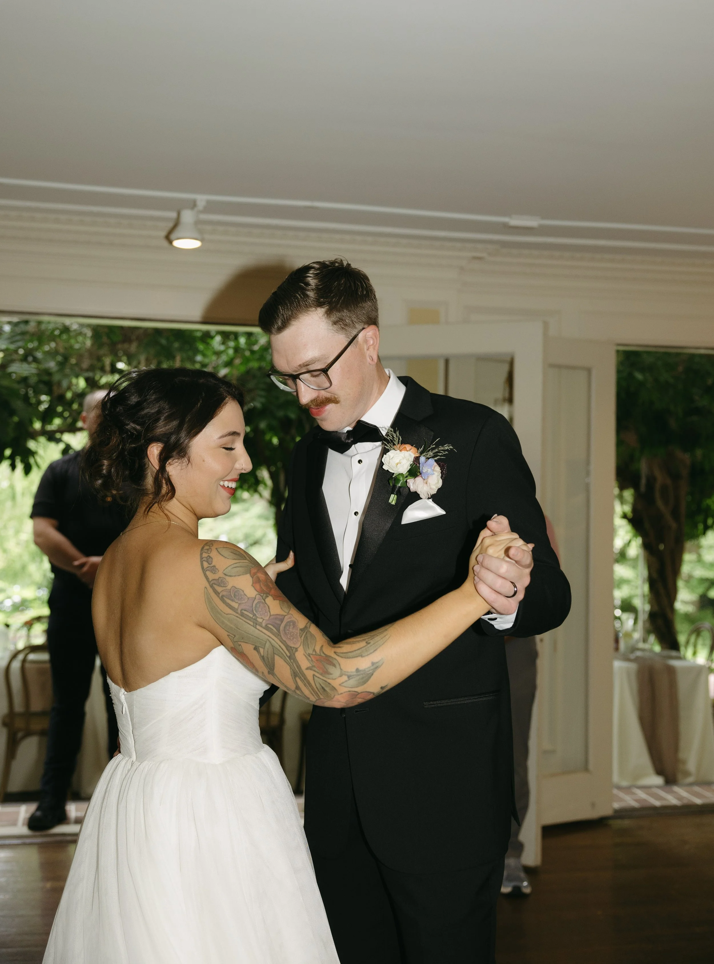 A couple dancing at their wedding, with the woman wearing a white strapless dress and the man in a black tuxedo, in a room with open doors and greenery outside.