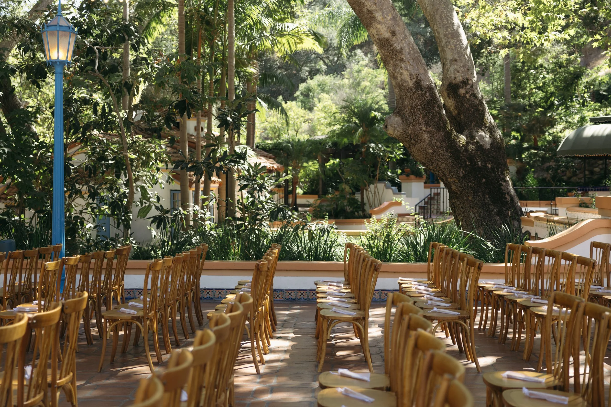 Outdoor ceremony area with wooden chairs, surrounded by lush greenery and large trees, in a sunny environment at a Rancho Las Lomas Wedding in Los Angeles