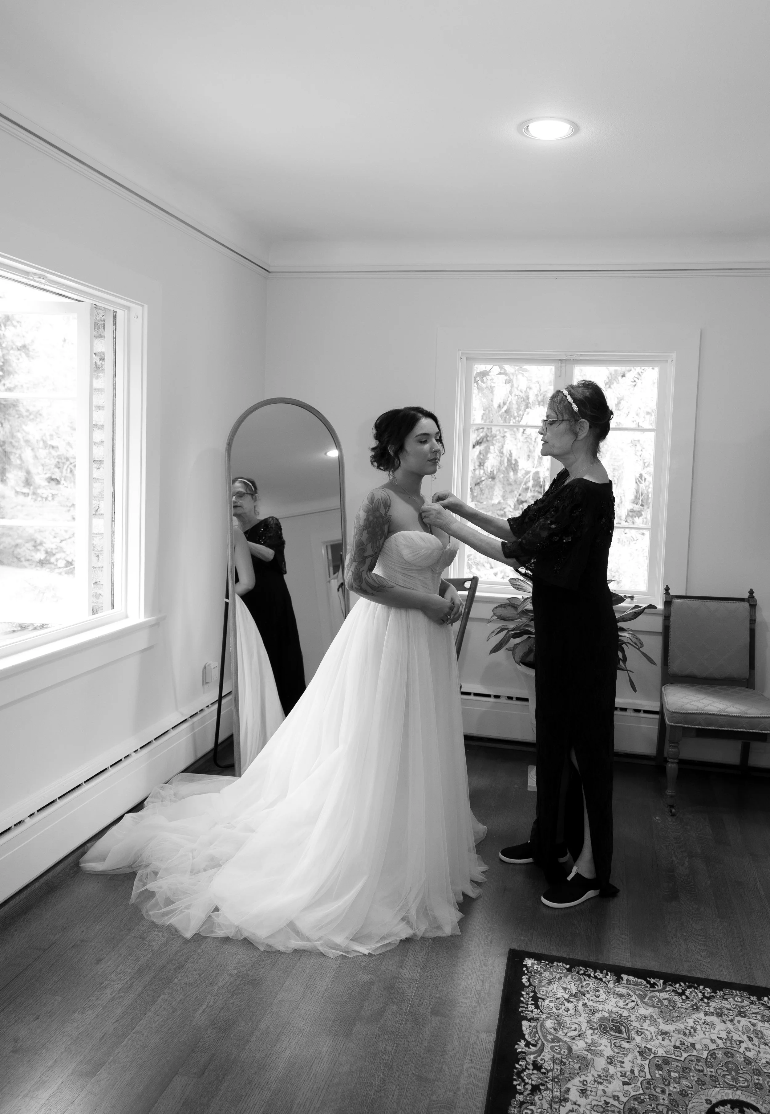 A bride getting ready in a bedroom as her mother adjusts her wedding dress. A mirror reflects the scene, and there are two windows providing natural light at a European inspired Lakewold Gardens wedding in Seattle