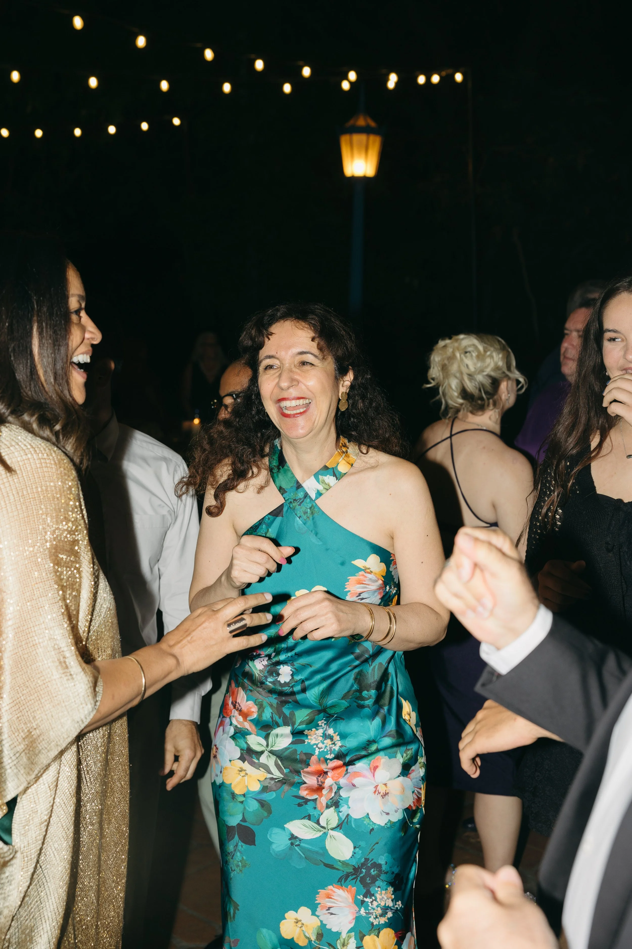 Group of people at a celebration, enjoying conversation and dancing. The scene is outdoors with string lights and a lamp post overhead.