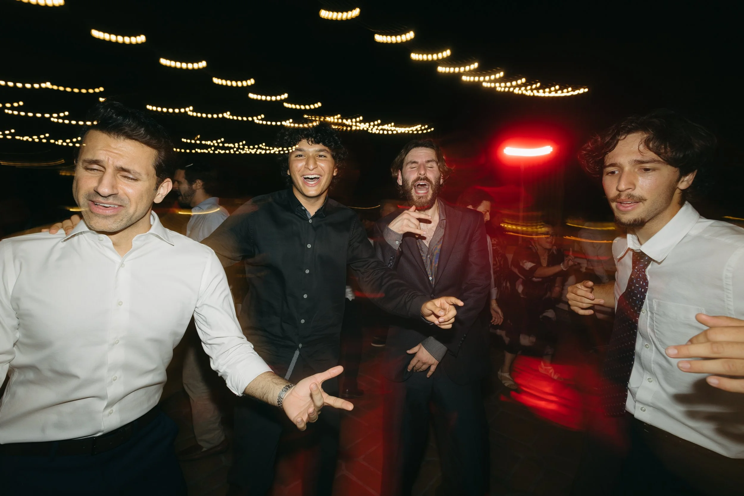 Group of four men dancing and enjoying a party at night with string lights overhead.