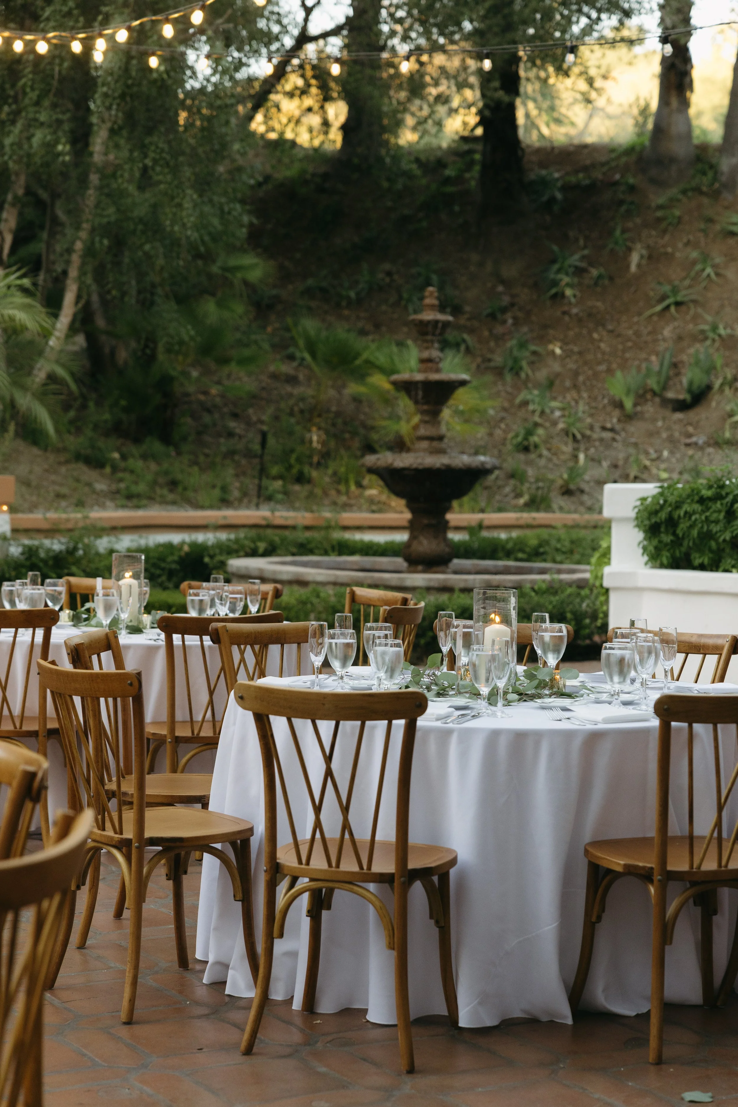 Outdoor reception dinner space, decorated with glasses, plates, silverware, candles, and greenery, with a multi-tiered fountain and string lights in the background at a Rancho Las Lomas Wedding in Los Angeles