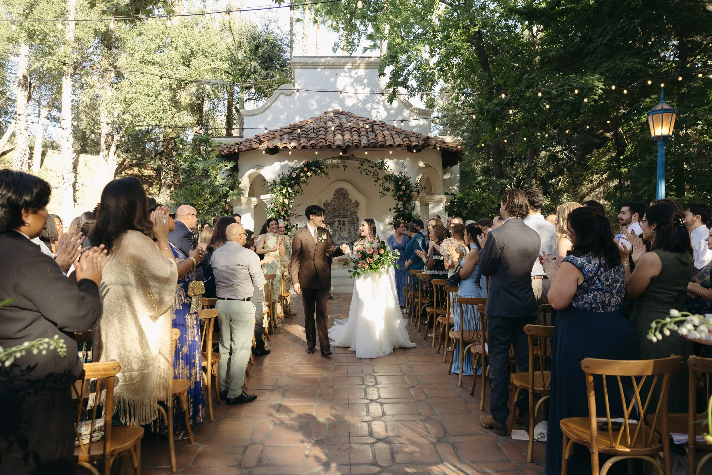 A wedding ceremony taking place outdoors with guests standing and clapping, a bride and groom walking down the aisle, surrounded by trees and string lights, at a Rancho Las Lomas Wedding in Los Angeles
