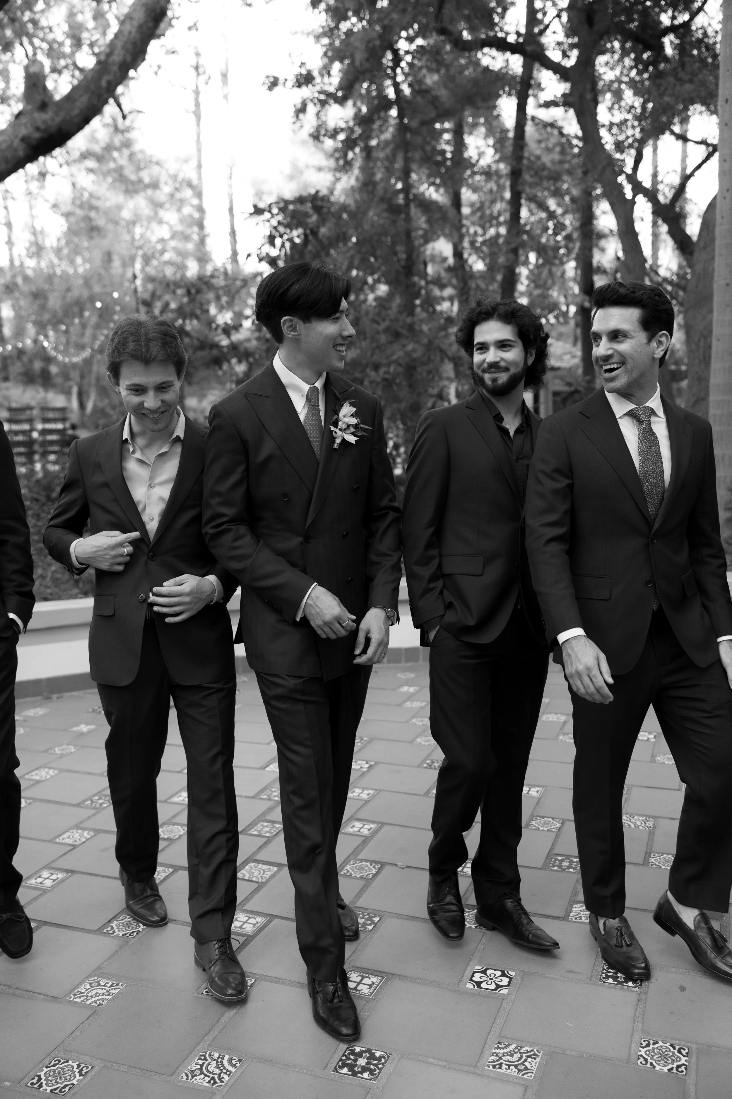 Black and white photo of four men dressed in suits, walking outdoors on a tiled patio surrounded by trees, smiling and chatting.