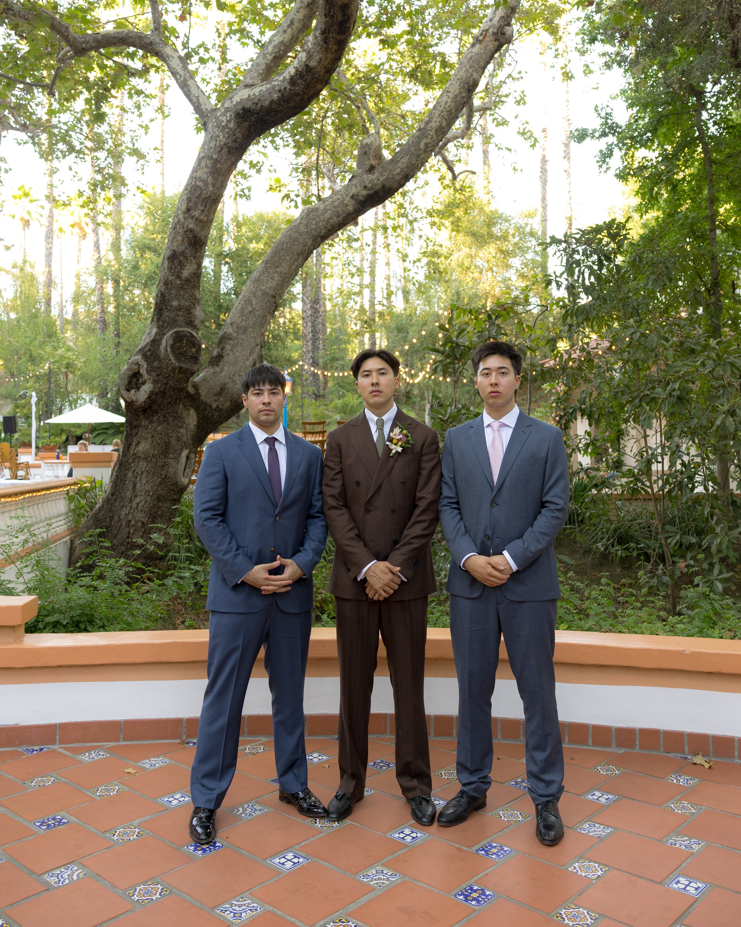 Three men in suits standing on a tiled patio with a large tree and greenery in the background, during a formal outdoor event.