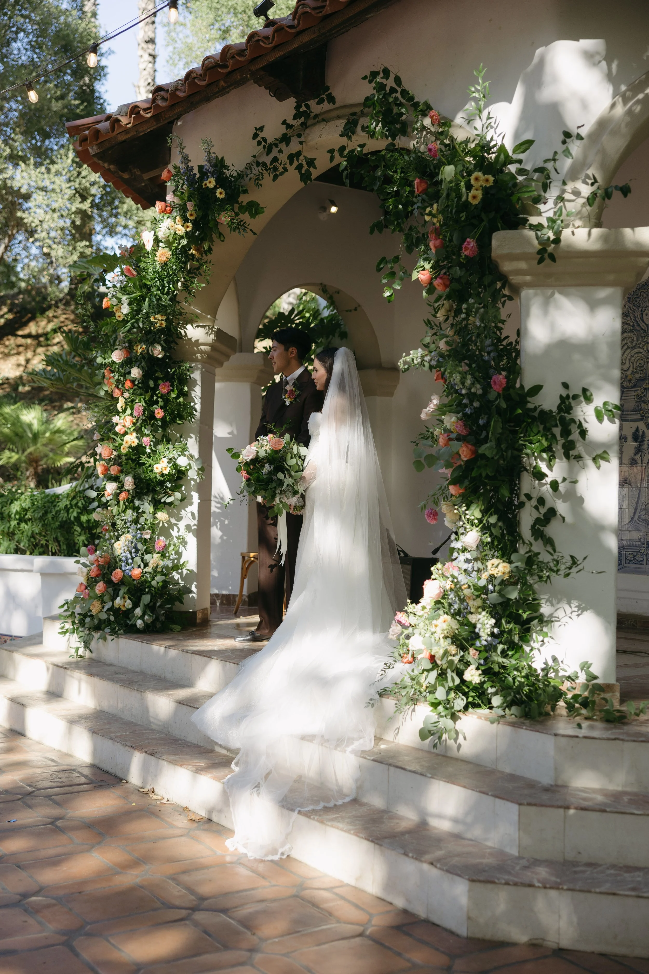 A bride and groom stand under a floral arch during their outdoor wedding ceremony, with trees and greenery in the background.