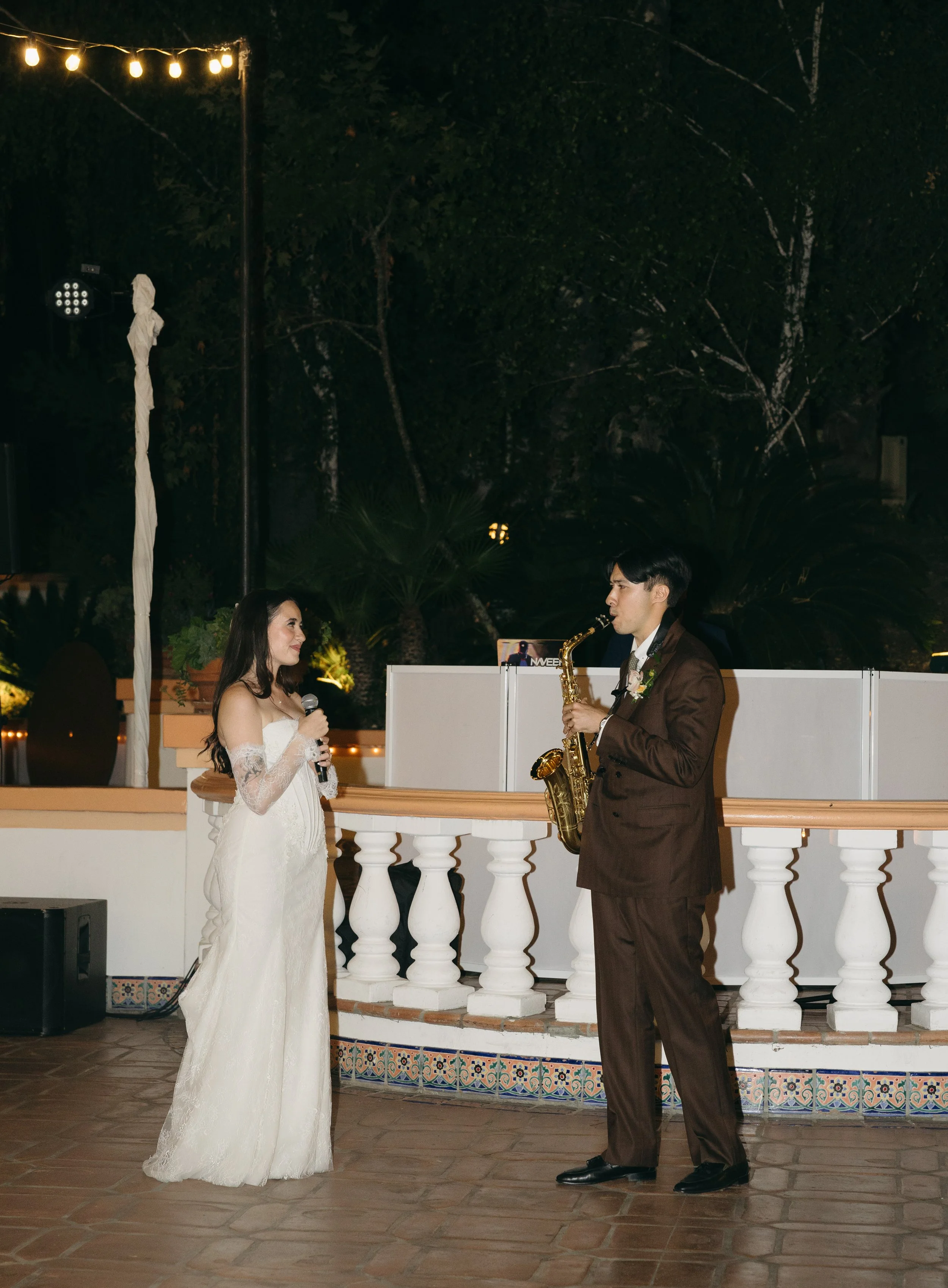 A bride and groom at their wedding reception, with the bride holding a microphone and the groom playing a saxophone, standing on a decorated outdoor patio at night.