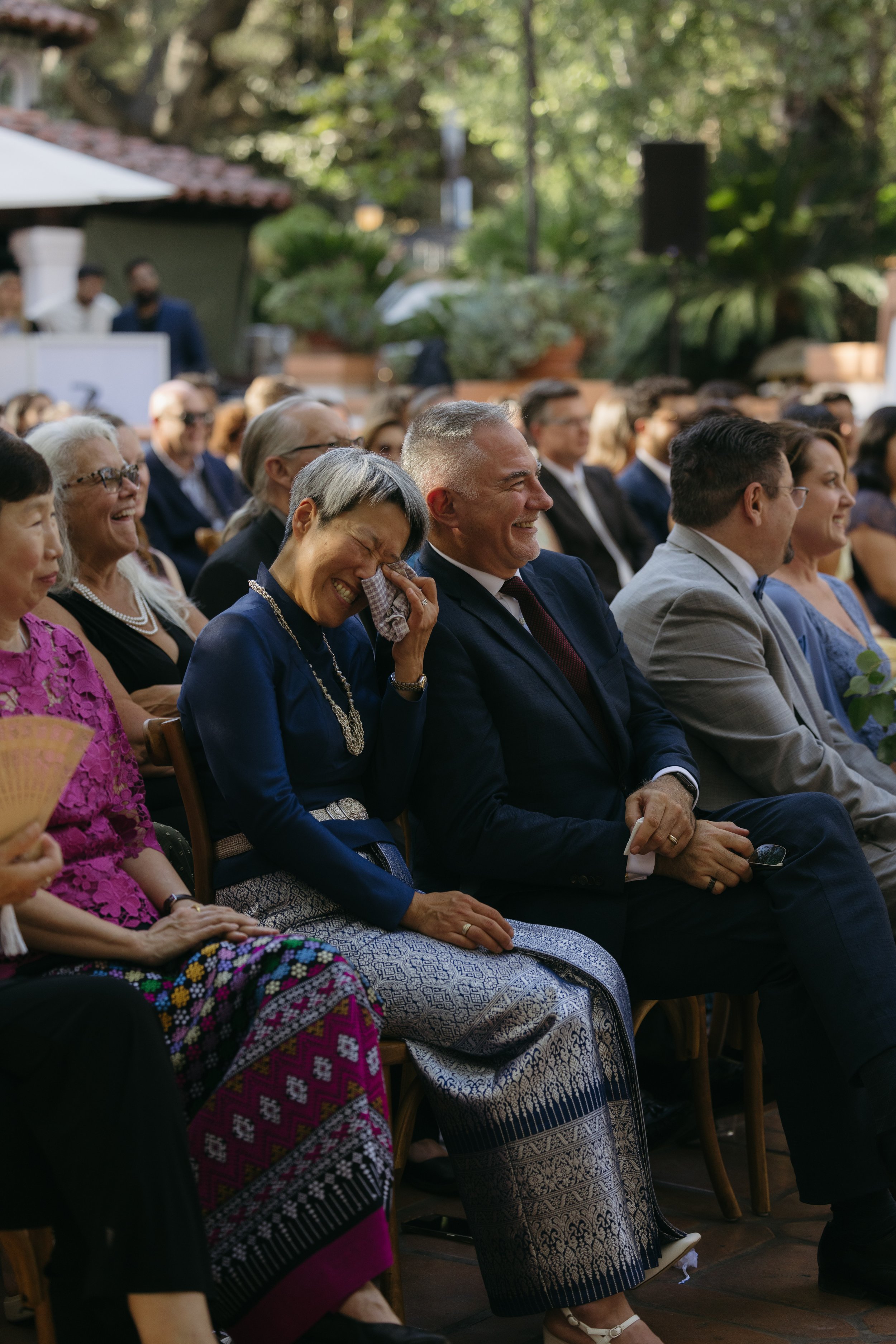 People attending a formal outdoor event, sitting on chairs, dressed in suits and traditional clothing, smiling and enjoying the moment.