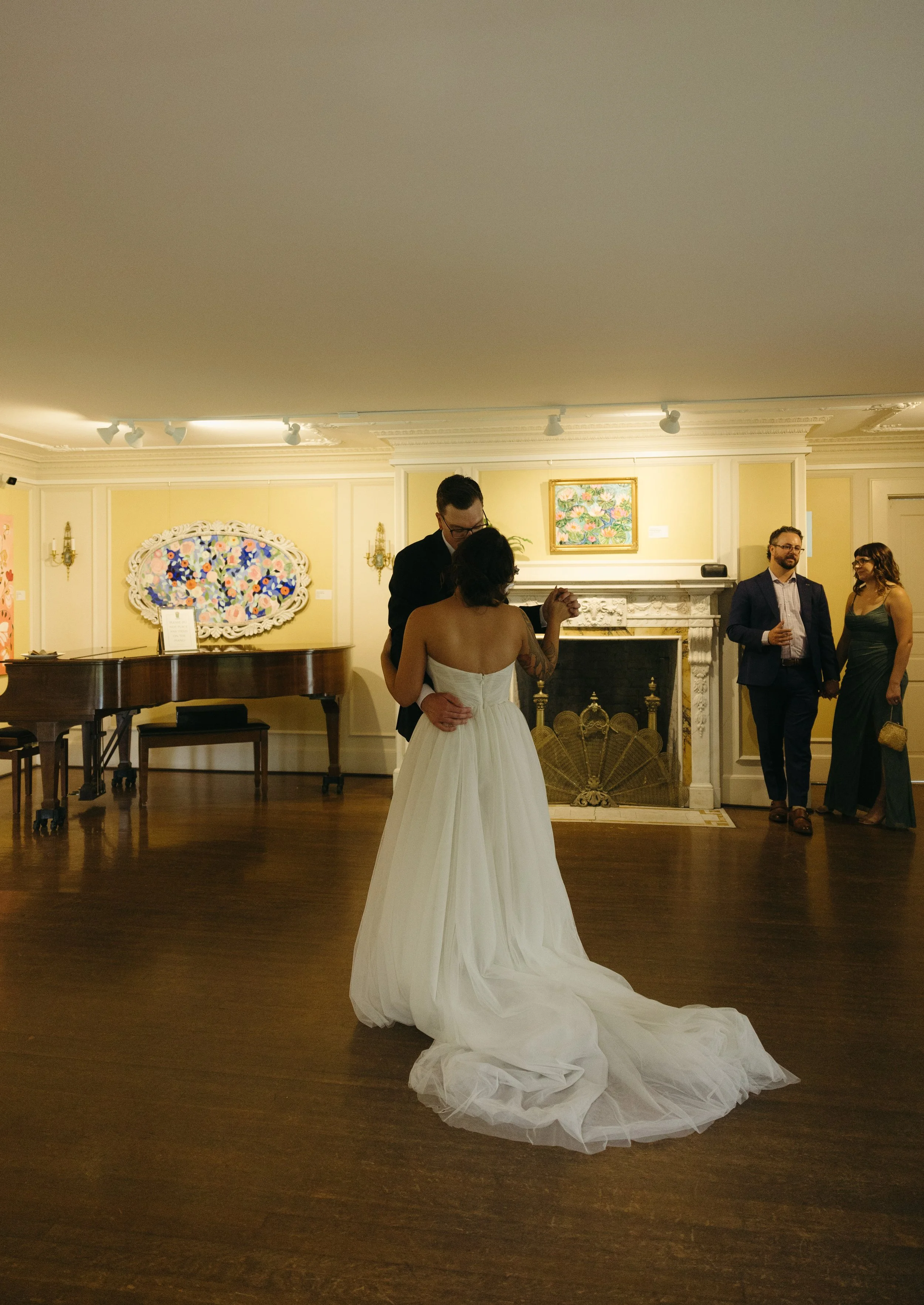 A bride and groom dancing in a warmly lit room with a fireplace, a piano, and guests watching.