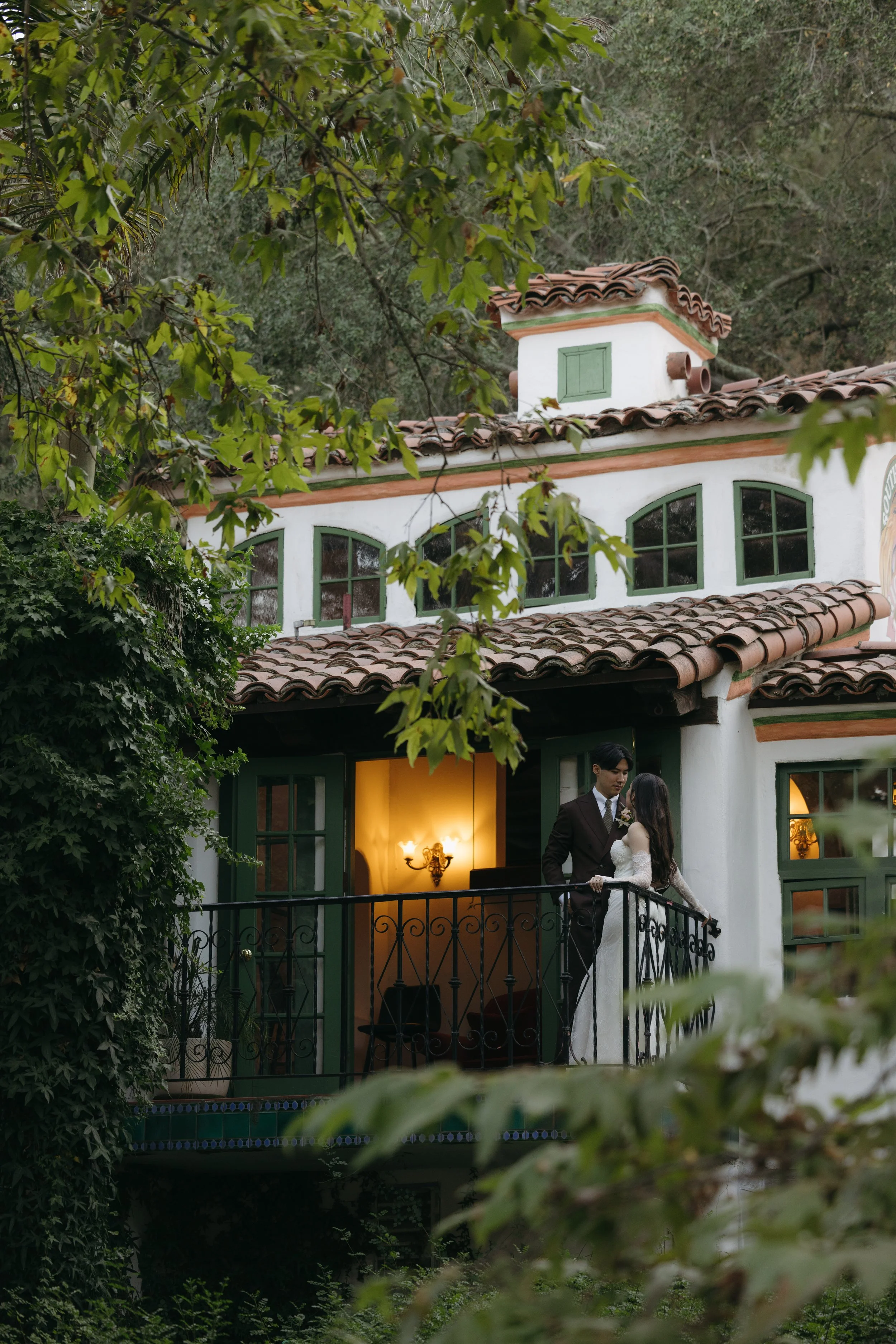 A couple dressed in formal attire standing on a balcony of a Spanish-style house surrounded by trees.